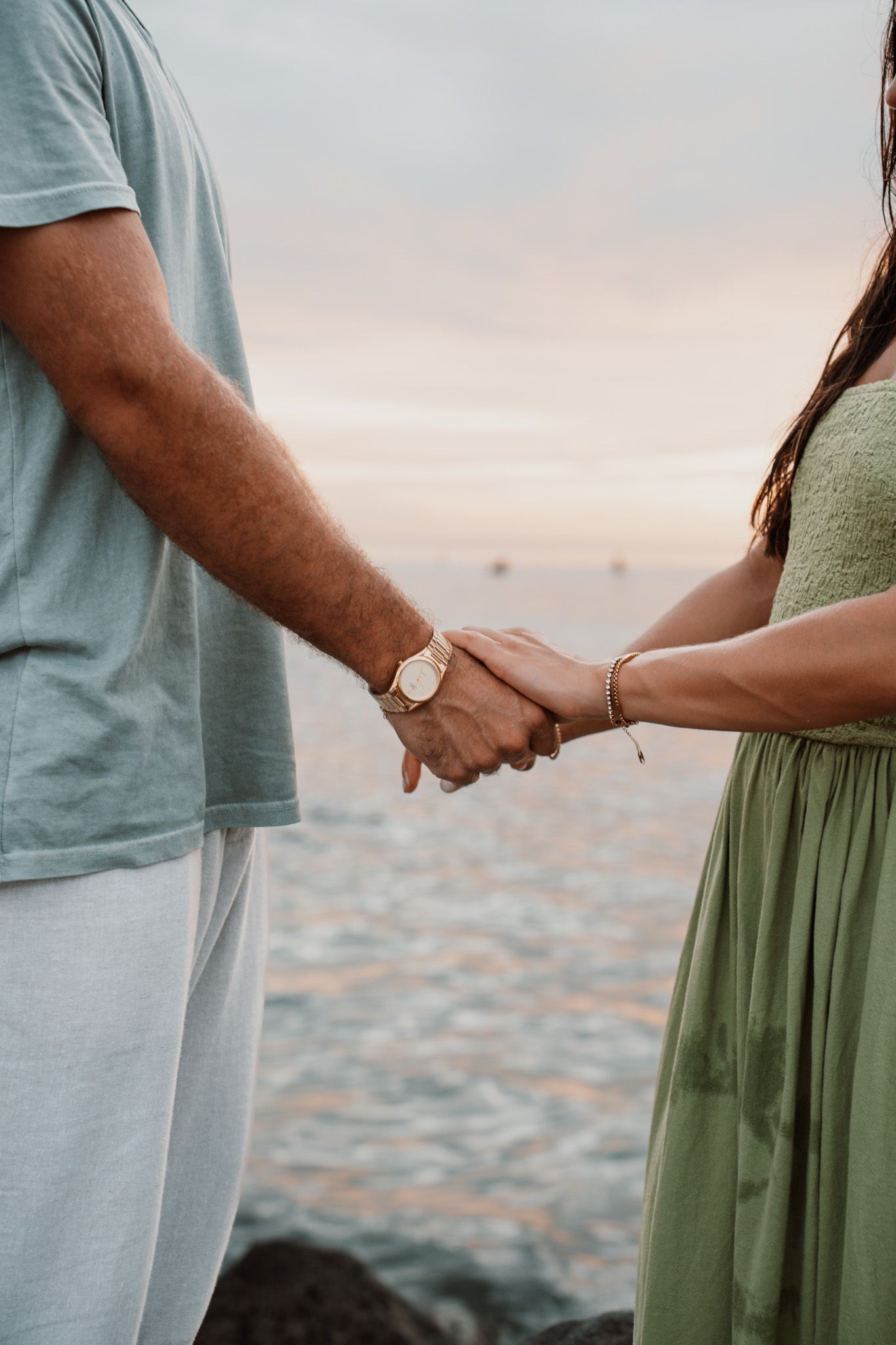 Couple holding hands on a rocky beach at sunset, man wearing a watch, woman in a green dress with a gold bracelet.