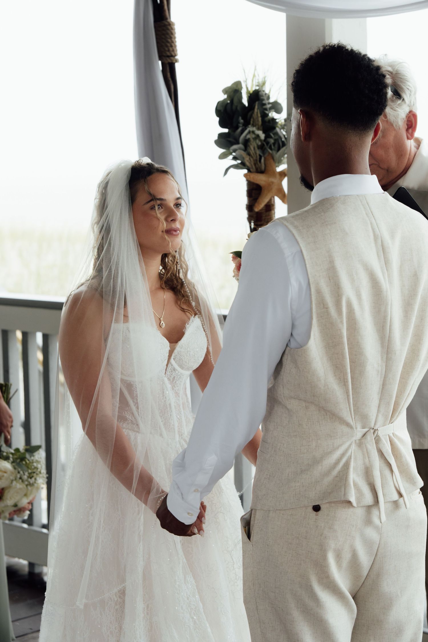 Bride in white gown and veil holds hands with groom in beige suit during outdoor wedding ceremony.