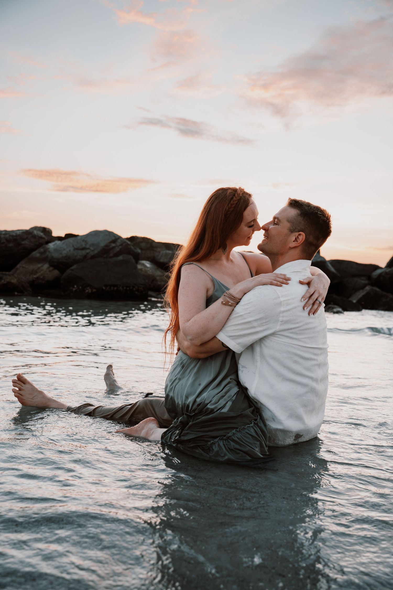 Couple embracing romantically in shallow ocean water at sunset, woman in green dress sitting on man's lap near rocky shore.