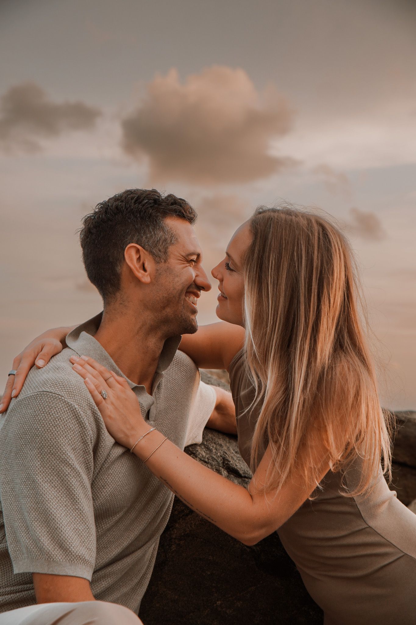 A smiling couple shares a romantic moment outdoors at sunset, with the woman leaning in close to the man.