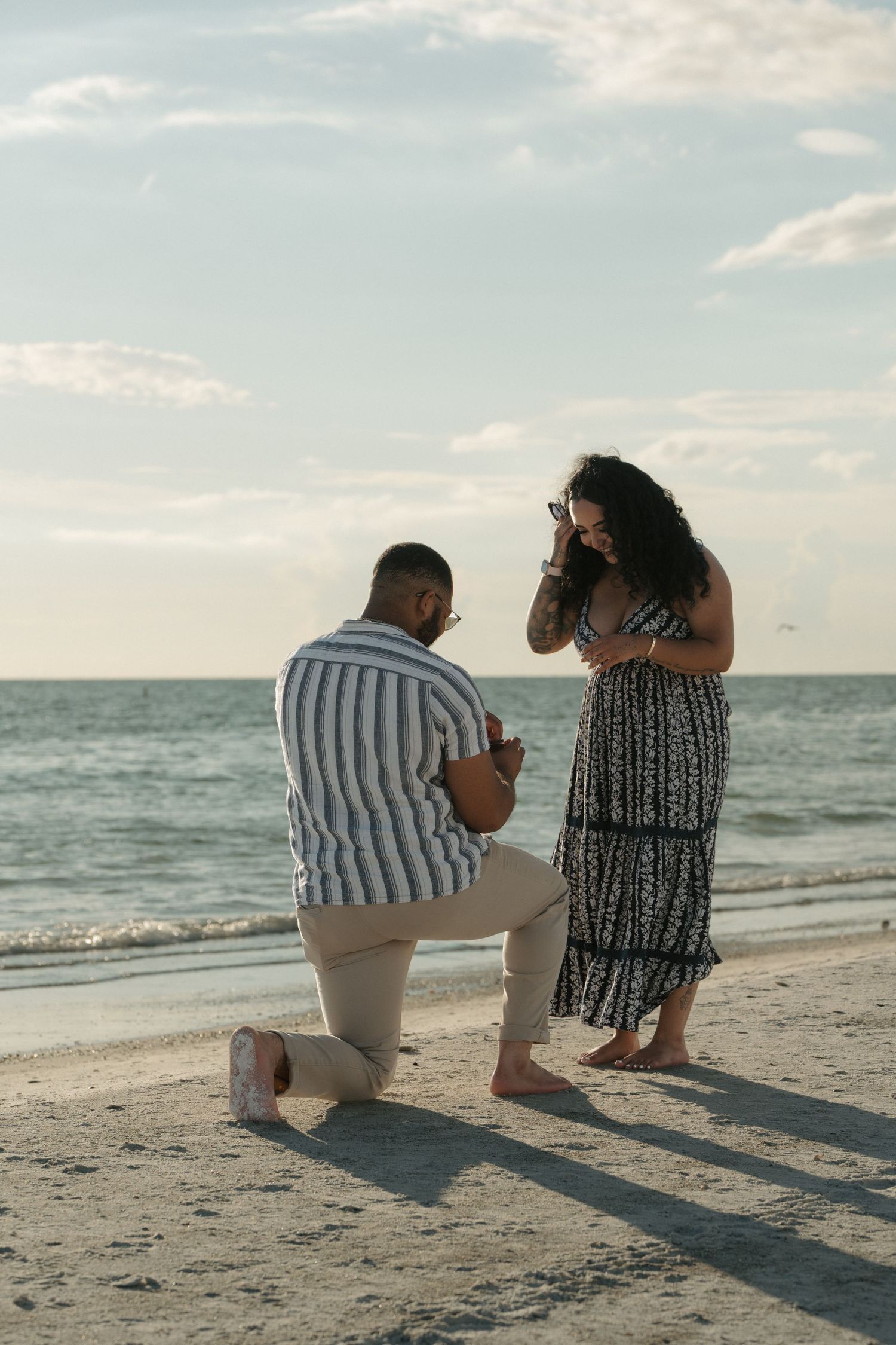 Man proposes on one knee to woman on a sandy beach at sunset, with ocean waves in the background.