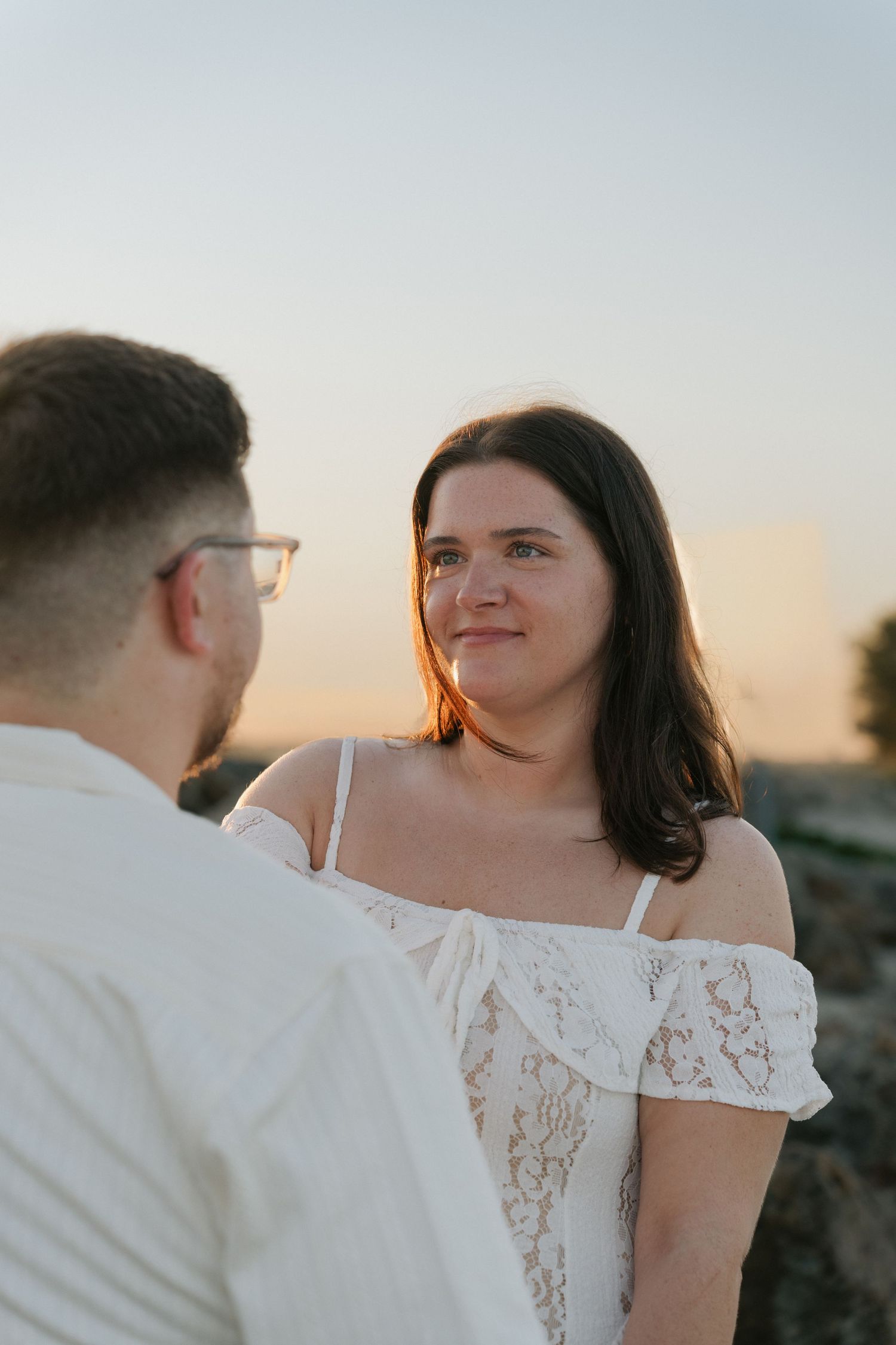 A bride in a white lace off-shoulder dress gazes lovingly at her groom during a golden hour outdoor ceremony.