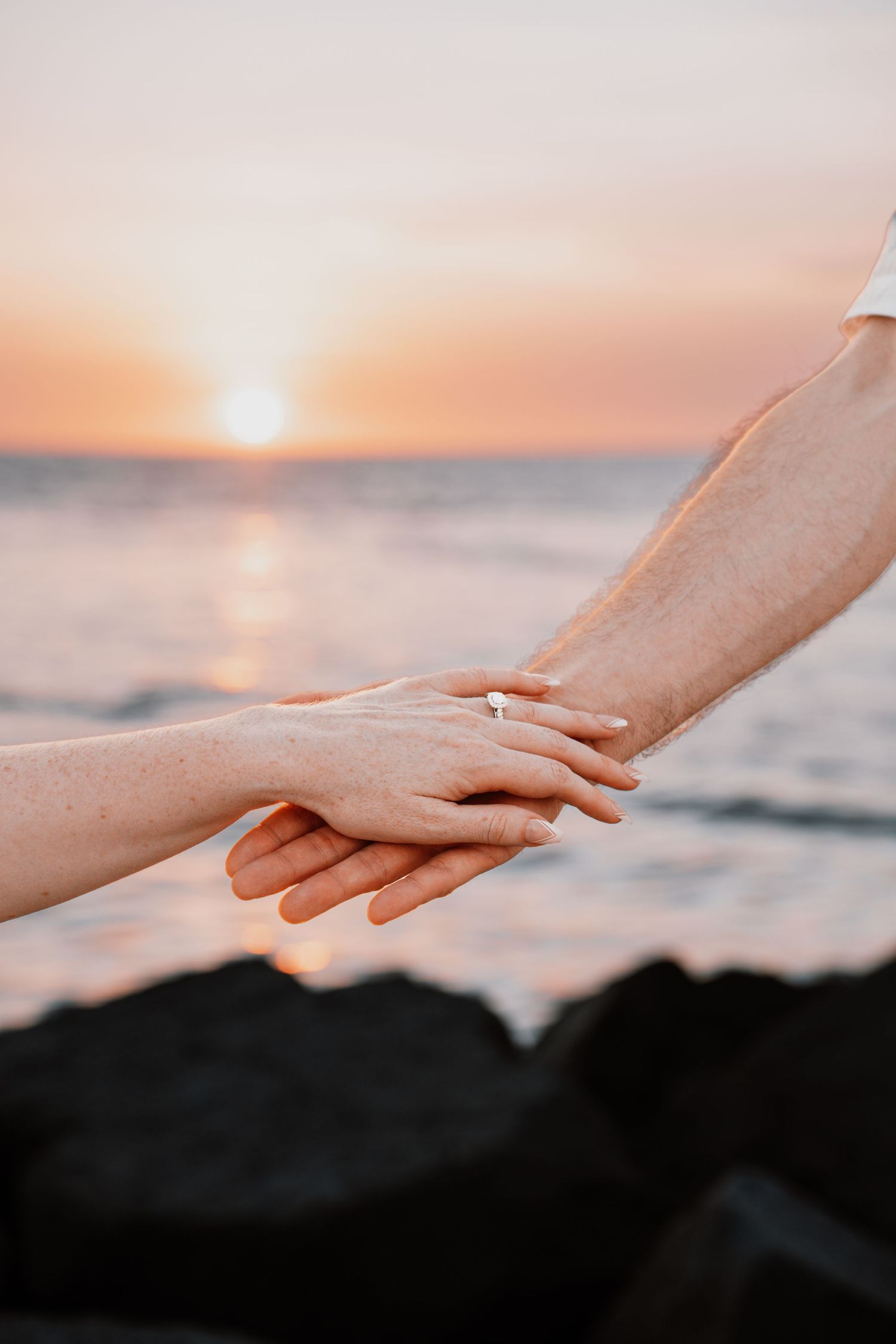 Couple holding hands with engagement ring at sunset by the ocean on rocky shore.