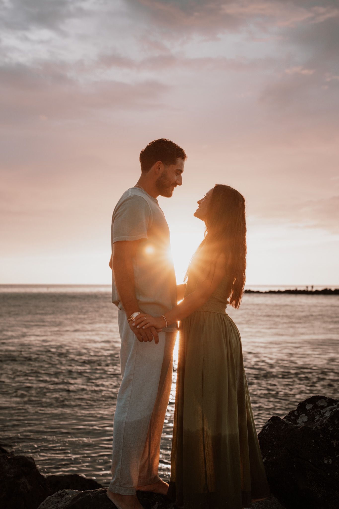 Romantic couple holding hands at sunset by the ocean, golden backlight glowing between them on rocky shore.