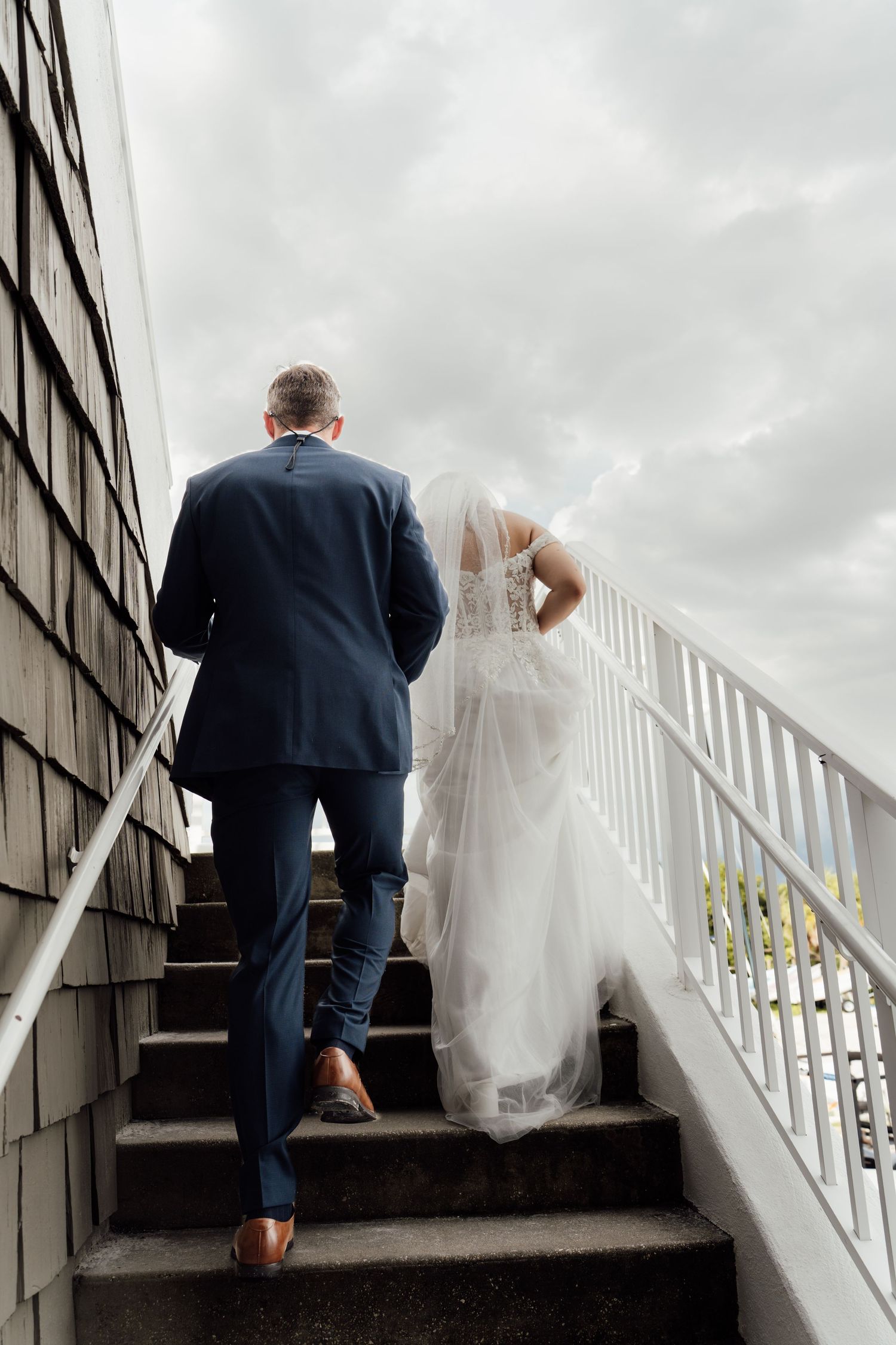 Bride and groom walking up outdoor stairs, bride in white gown with veil, groom in navy suit, cloudy sky backdrop.