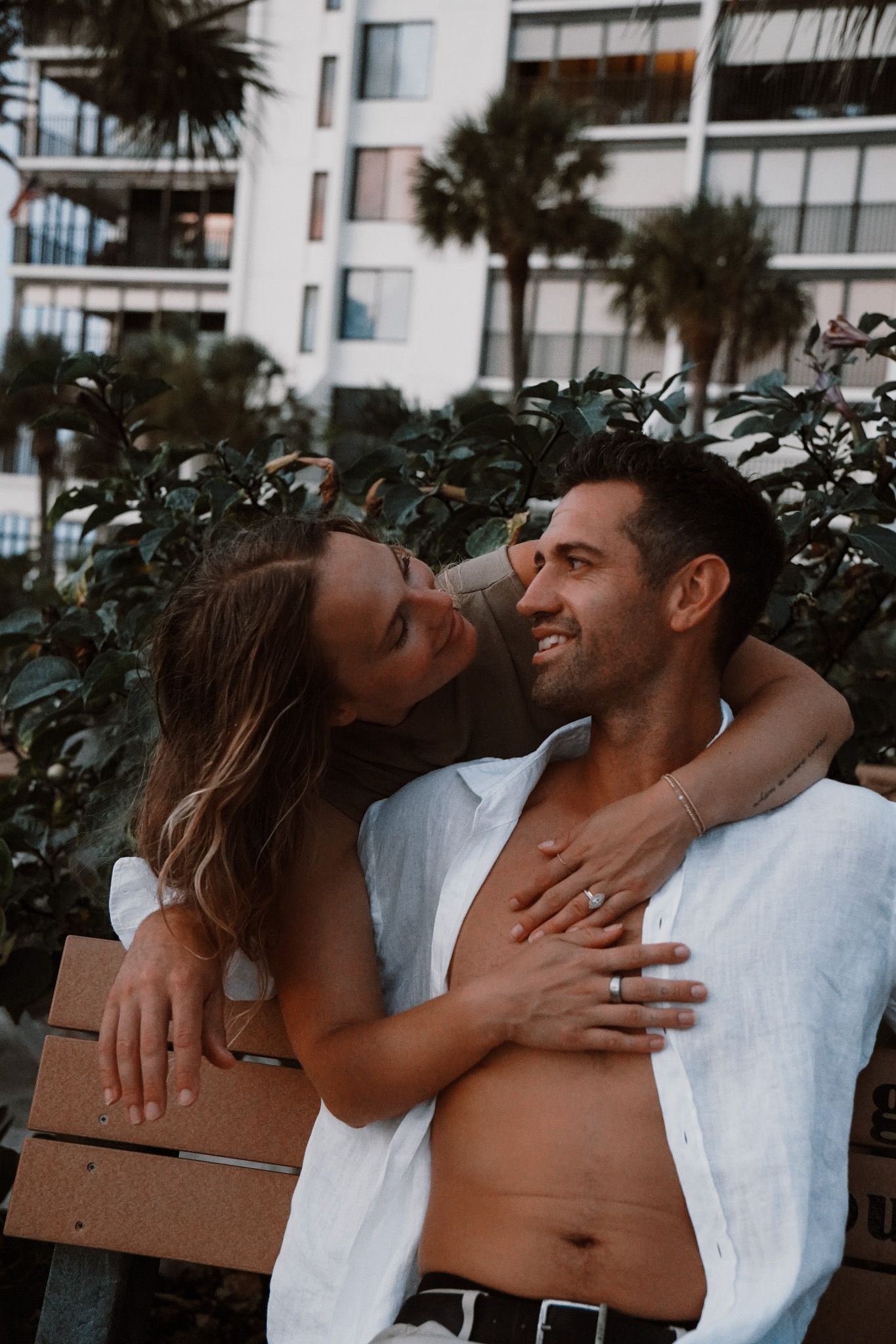 Couple embracing on a bench with palm trees and a building in the background, woman wearing an engagement ring.