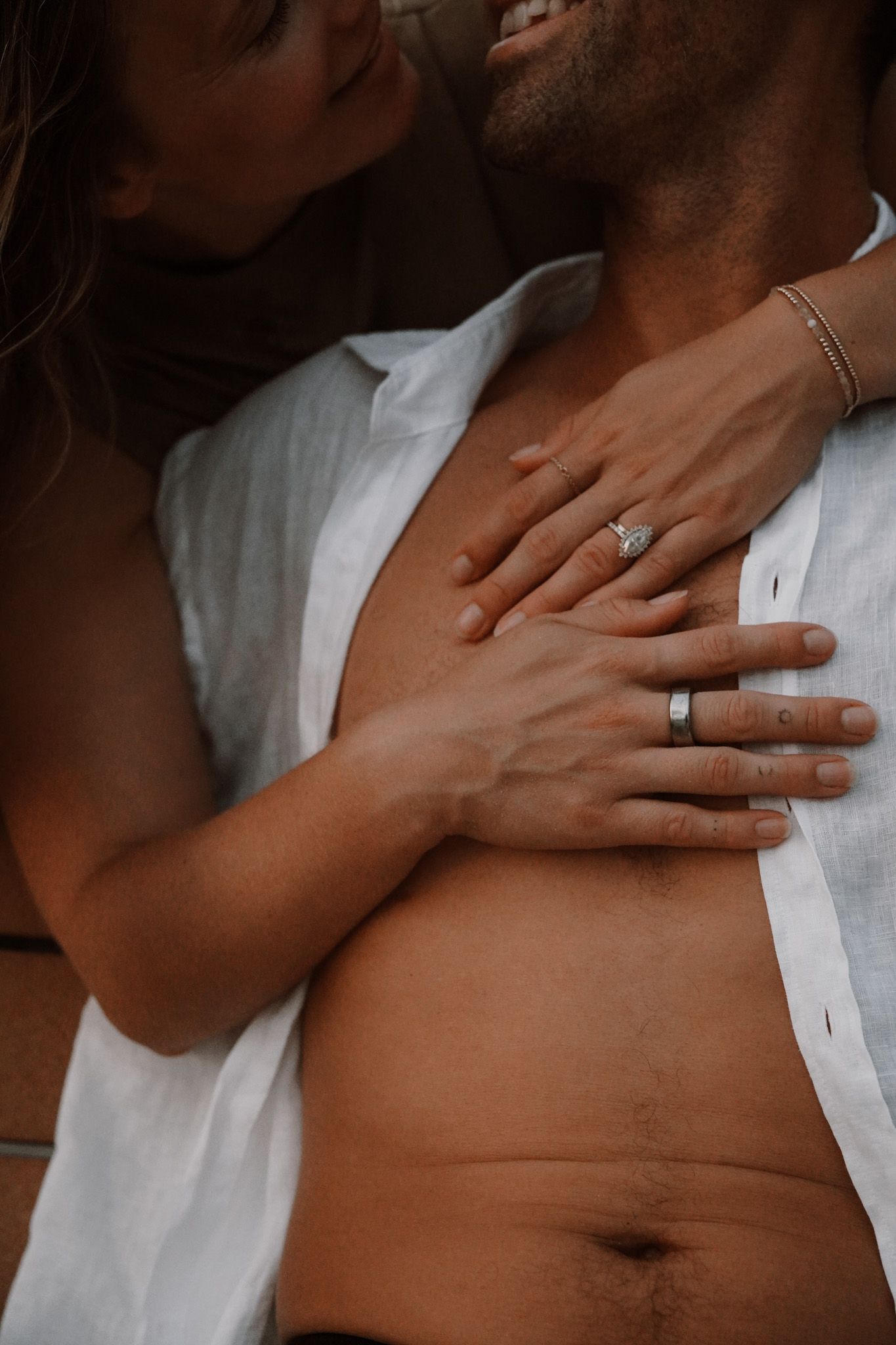 Couple embracing intimately, woman wearing engagement ring resting hands on man's chest in romantic pose.