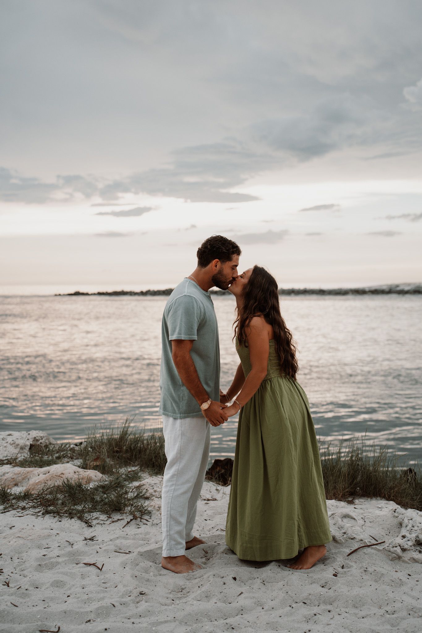 Couple kissing on a white sand beach at sunset, man in blue shirt and woman in green dress holding hands.