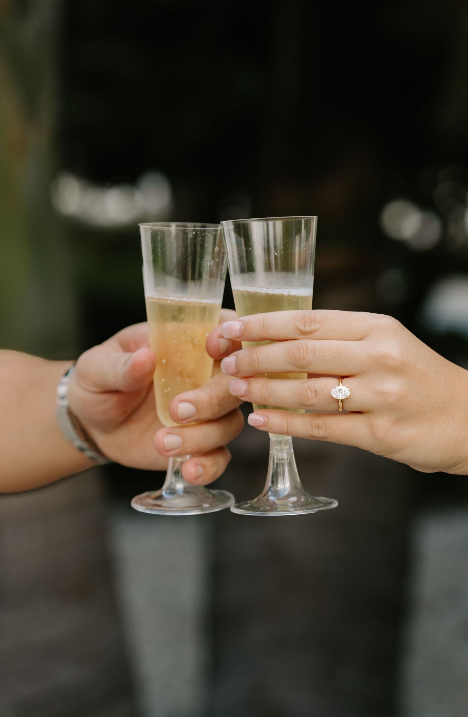 Two hands clinking champagne flutes in a toast, one adorned with a diamond engagement ring and the other with a silver bracelet.