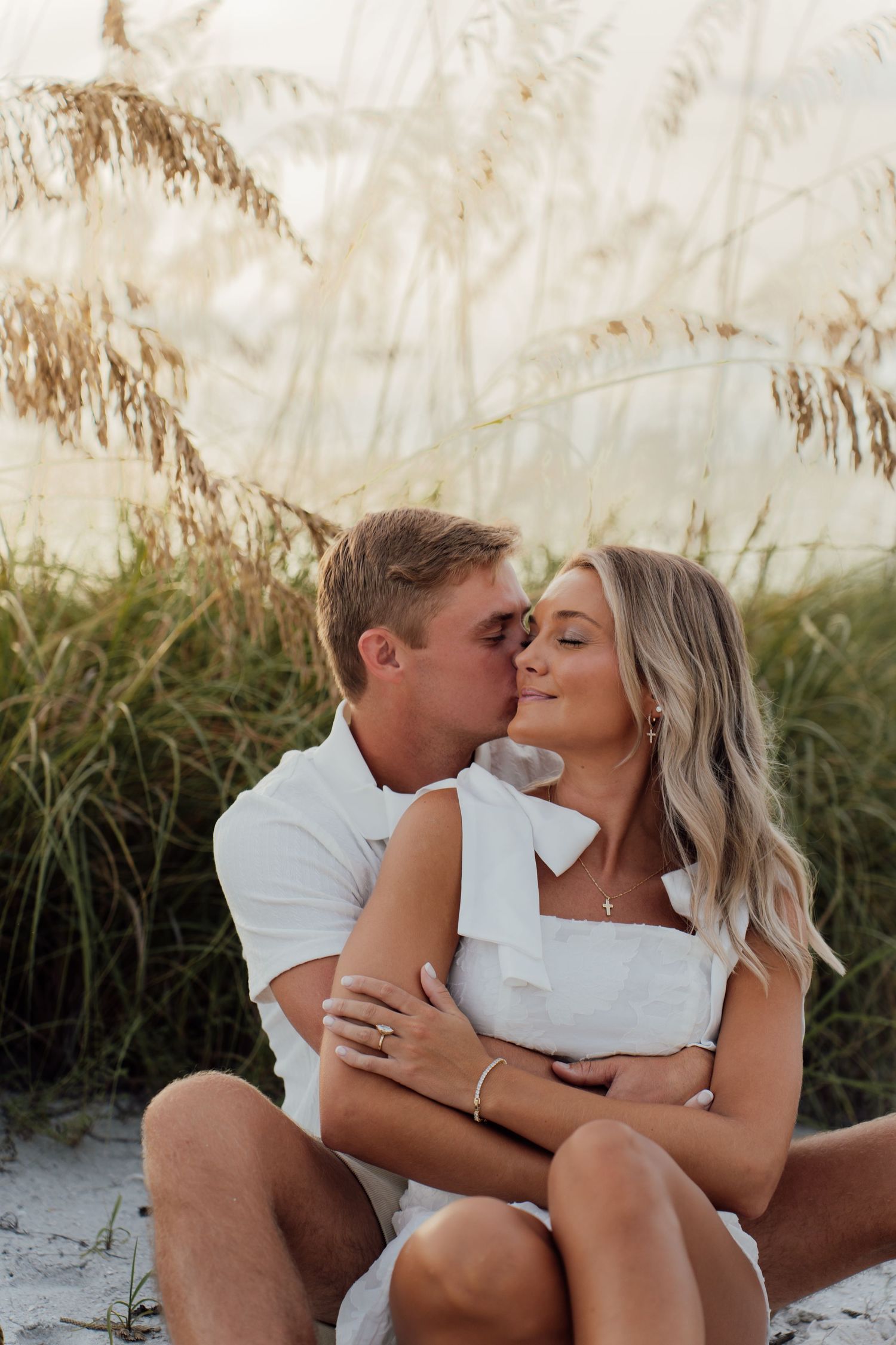 A couple shares a romantic kiss on a sandy beach surrounded by tall sea oats grass, both dressed in white.