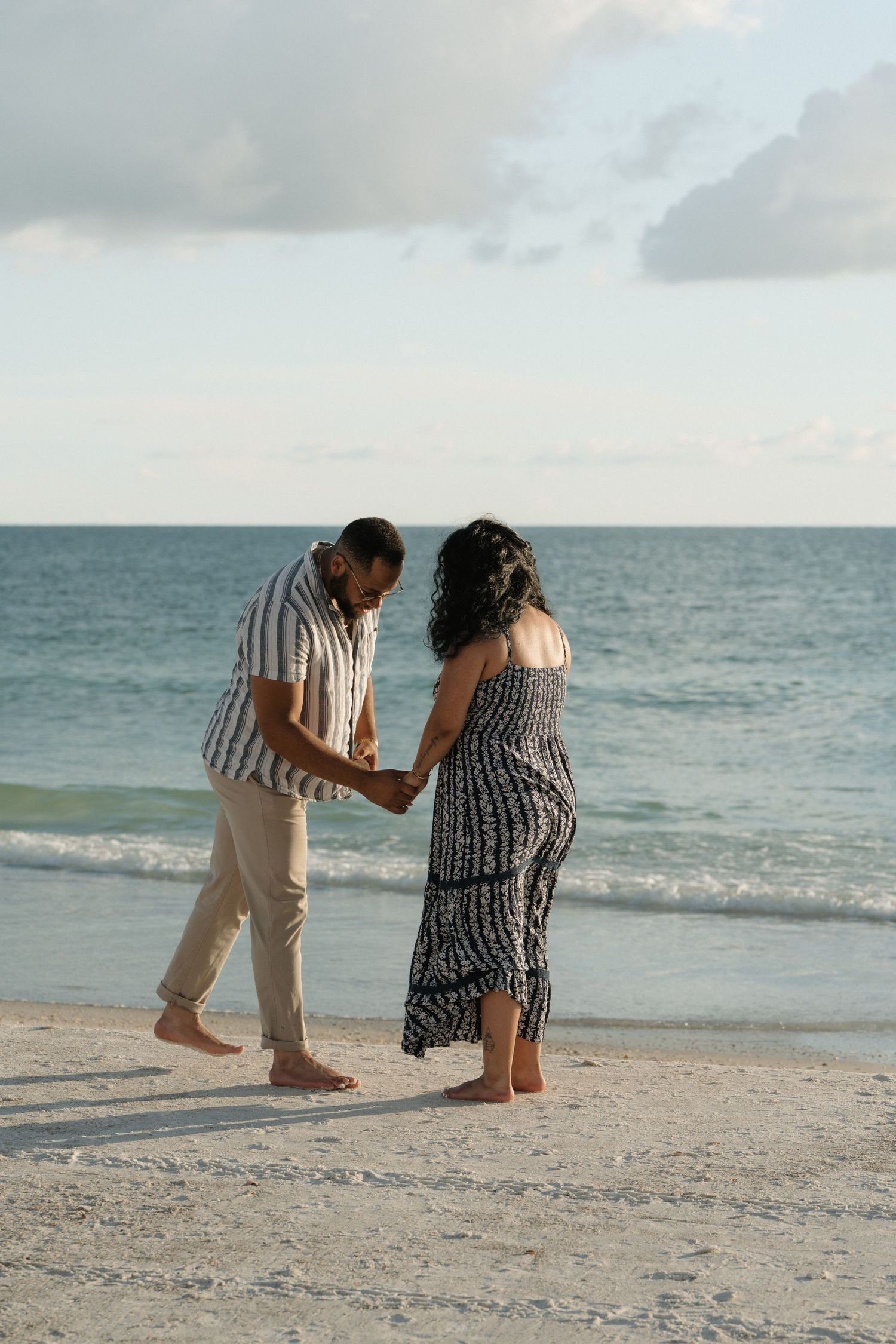Couple holding hands walking barefoot on a sandy beach with calm ocean waves in the background.