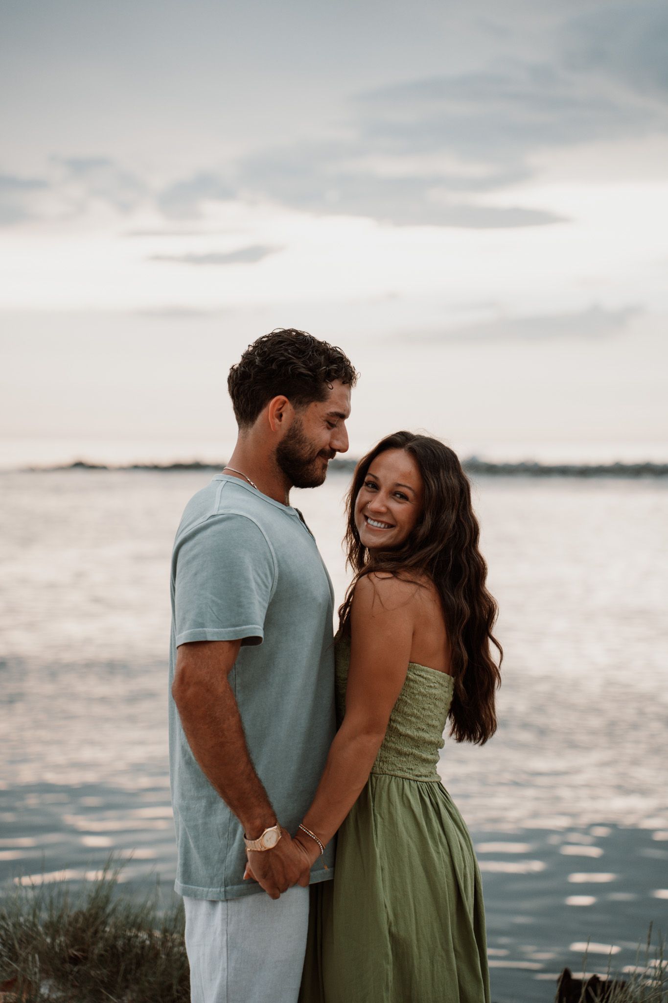 Couple holding hands and smiling near a waterfront at sunset, woman in green dress, man in light blue shirt.