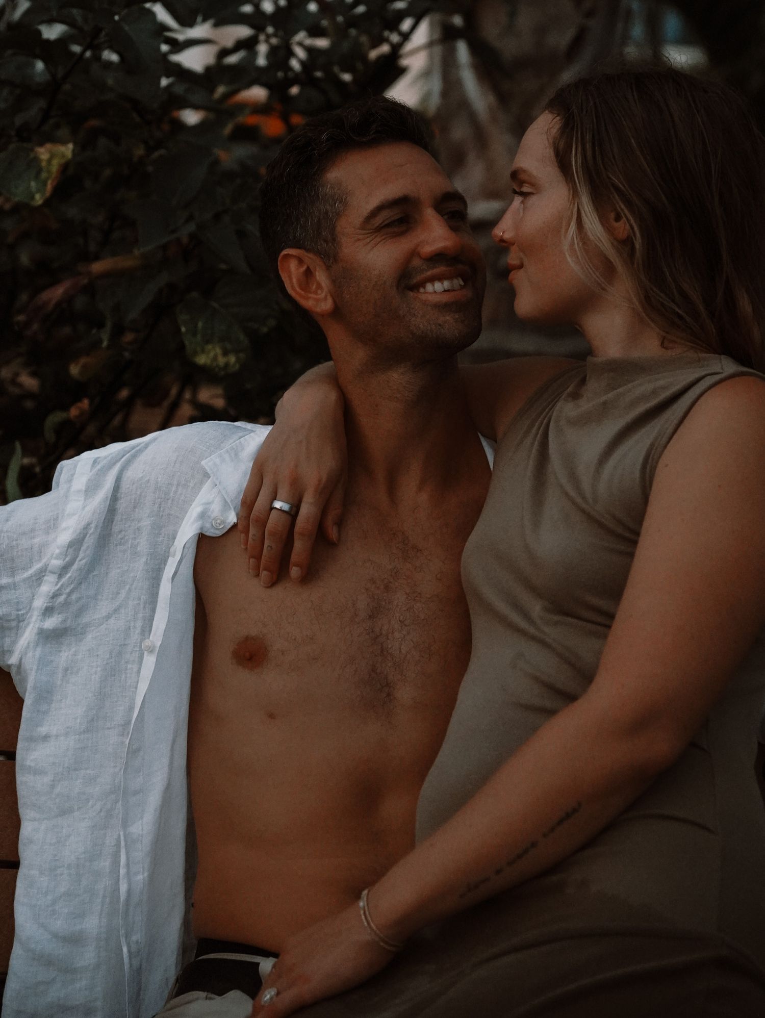 Smiling couple in an intimate outdoor moment, man shirtless with white shirt, woman in olive dress.