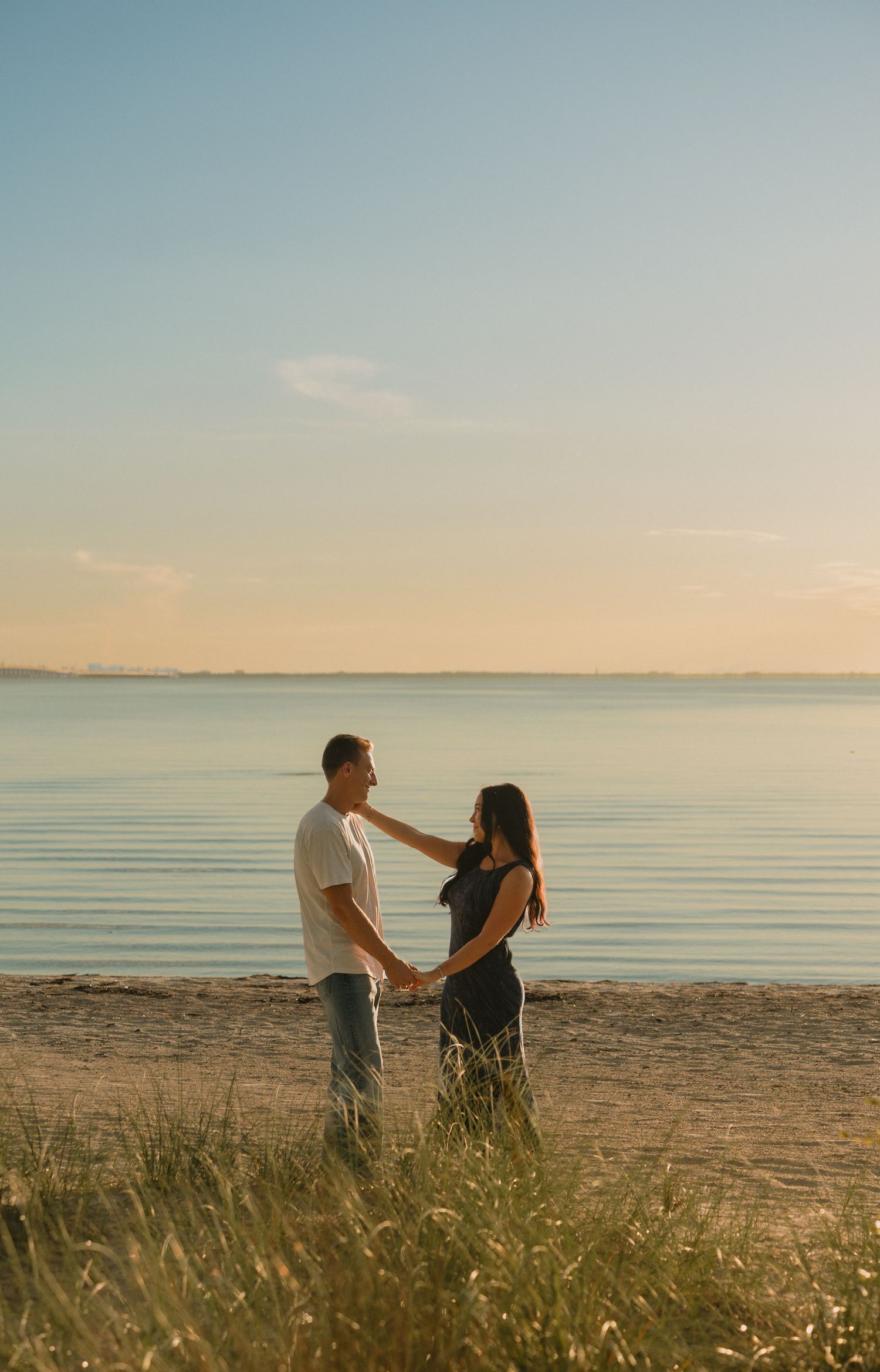 Couple holding hands on a sandy beach at sunset with calm water and golden sky in the background.