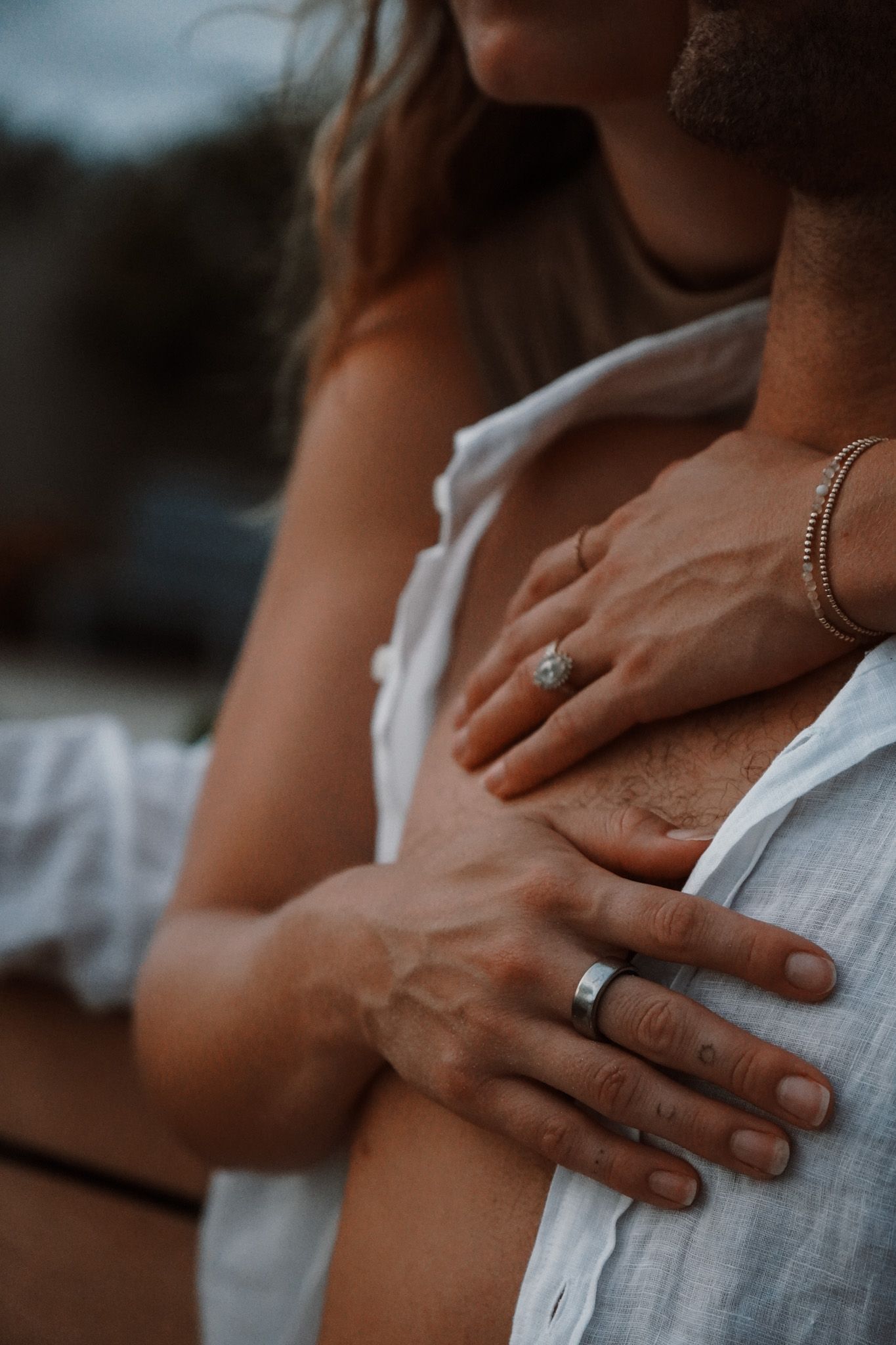 Close-up of couple embracing, woman wearing diamond ring and bracelet, man wearing silver band, both in white shirts.