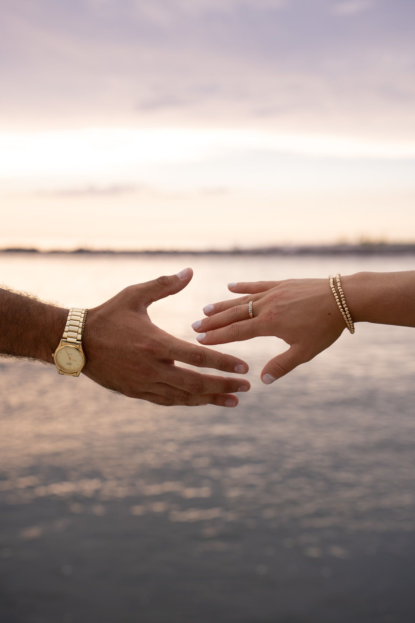 Two hands reaching toward each other at sunset, one wearing a gold watch, the other an engagement ring and bracelet.