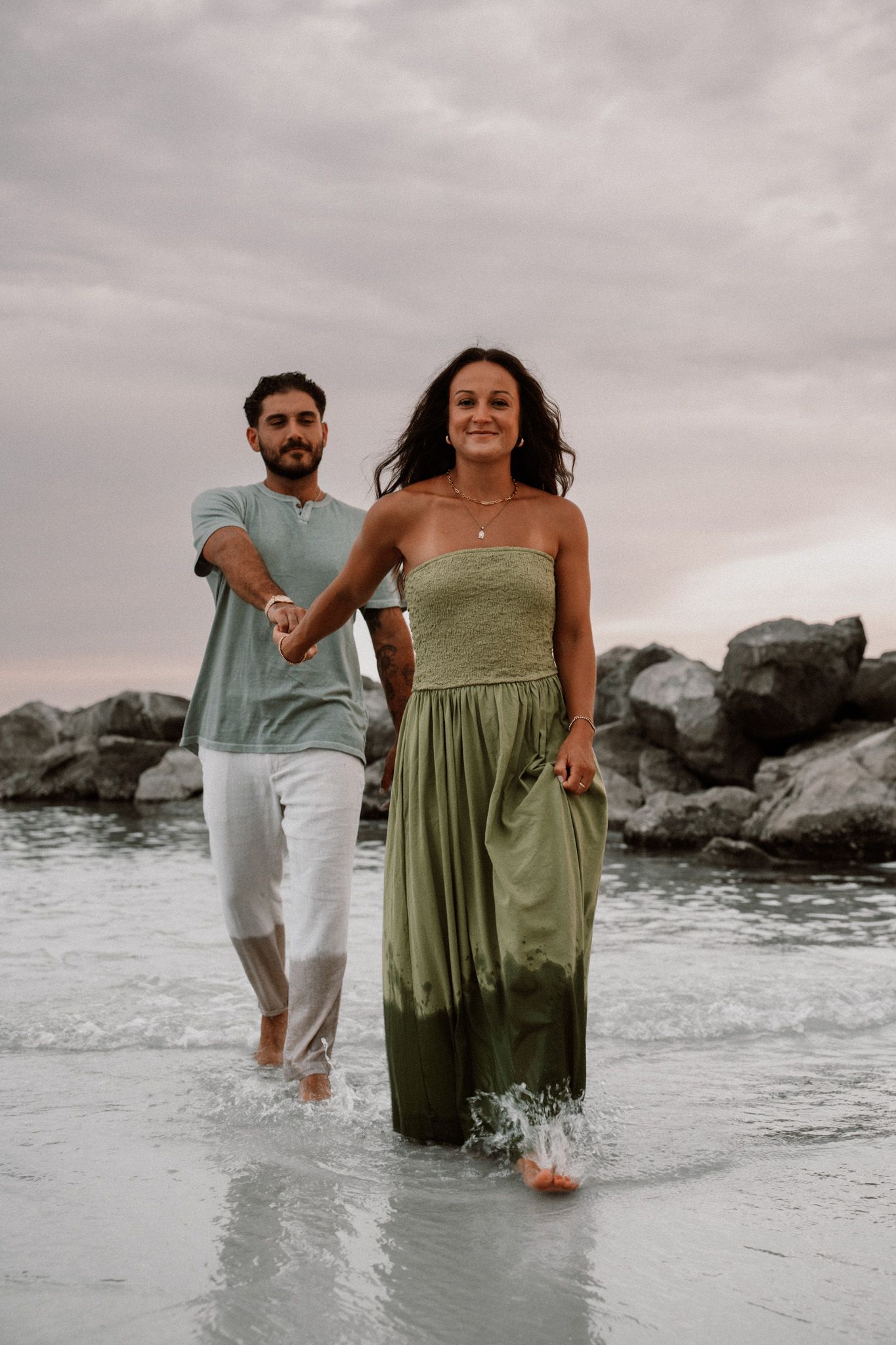 Couple walking hand-in-hand through ocean waves at sunset, woman in green maxi dress, man in teal shirt and white pants.