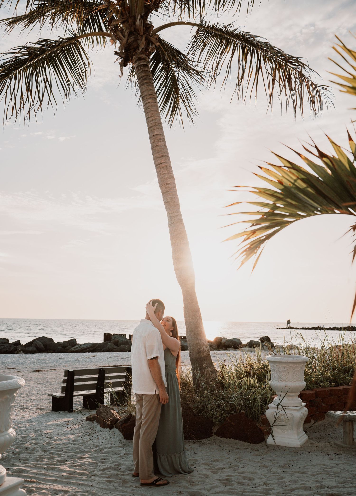 Couple embracing under a palm tree on a tropical beach at sunset with ocean views and lush greenery.