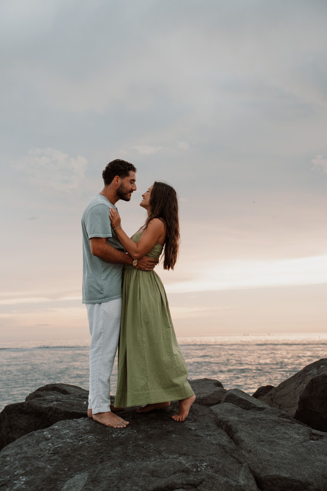 Couple embracing on rocky shore at sunset, woman in green maxi dress, man in light blue shirt and white pants.