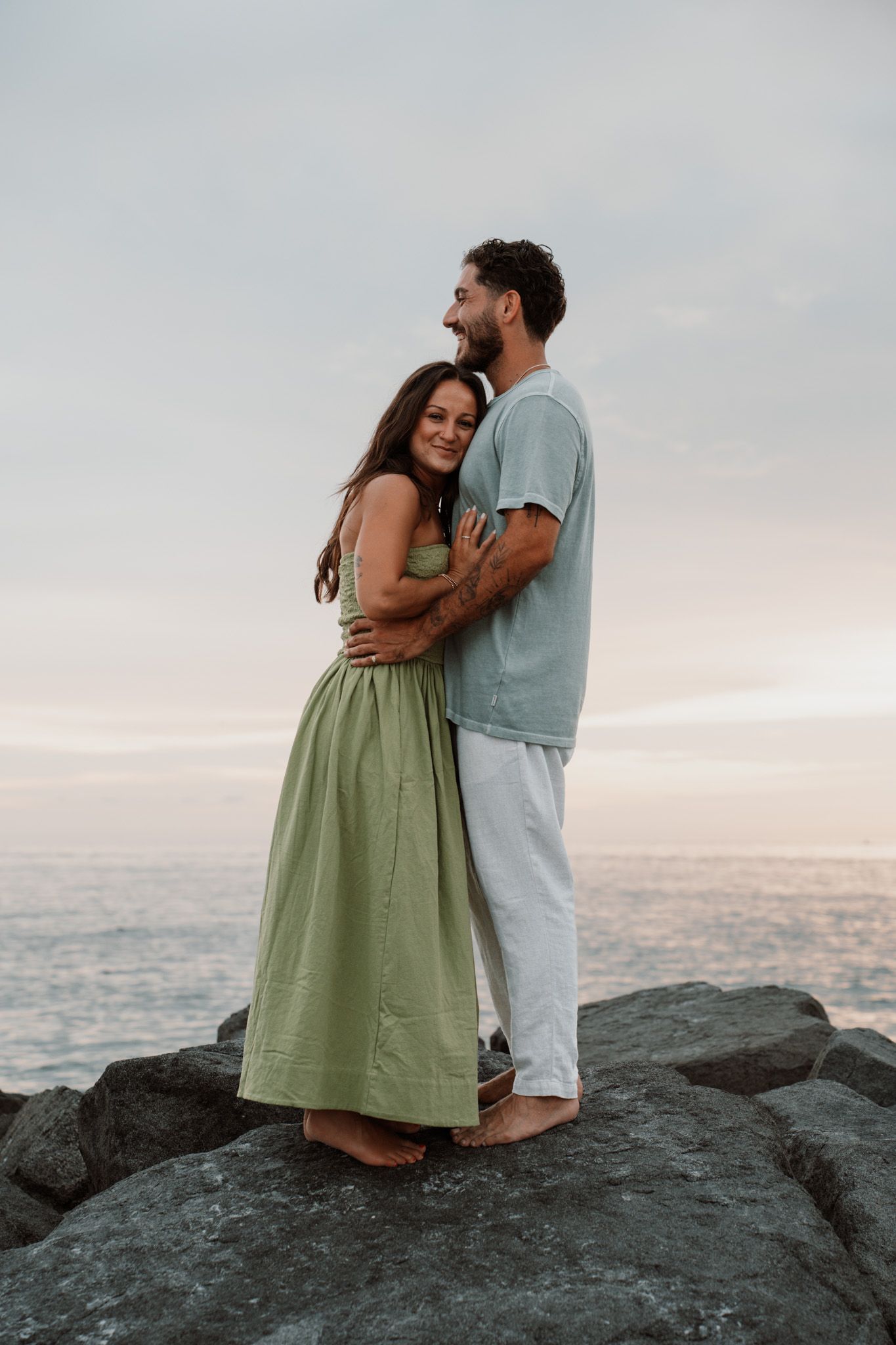Romantic couple embracing on rocky shoreline at sunset, woman in green dress leaning into man in teal shirt.