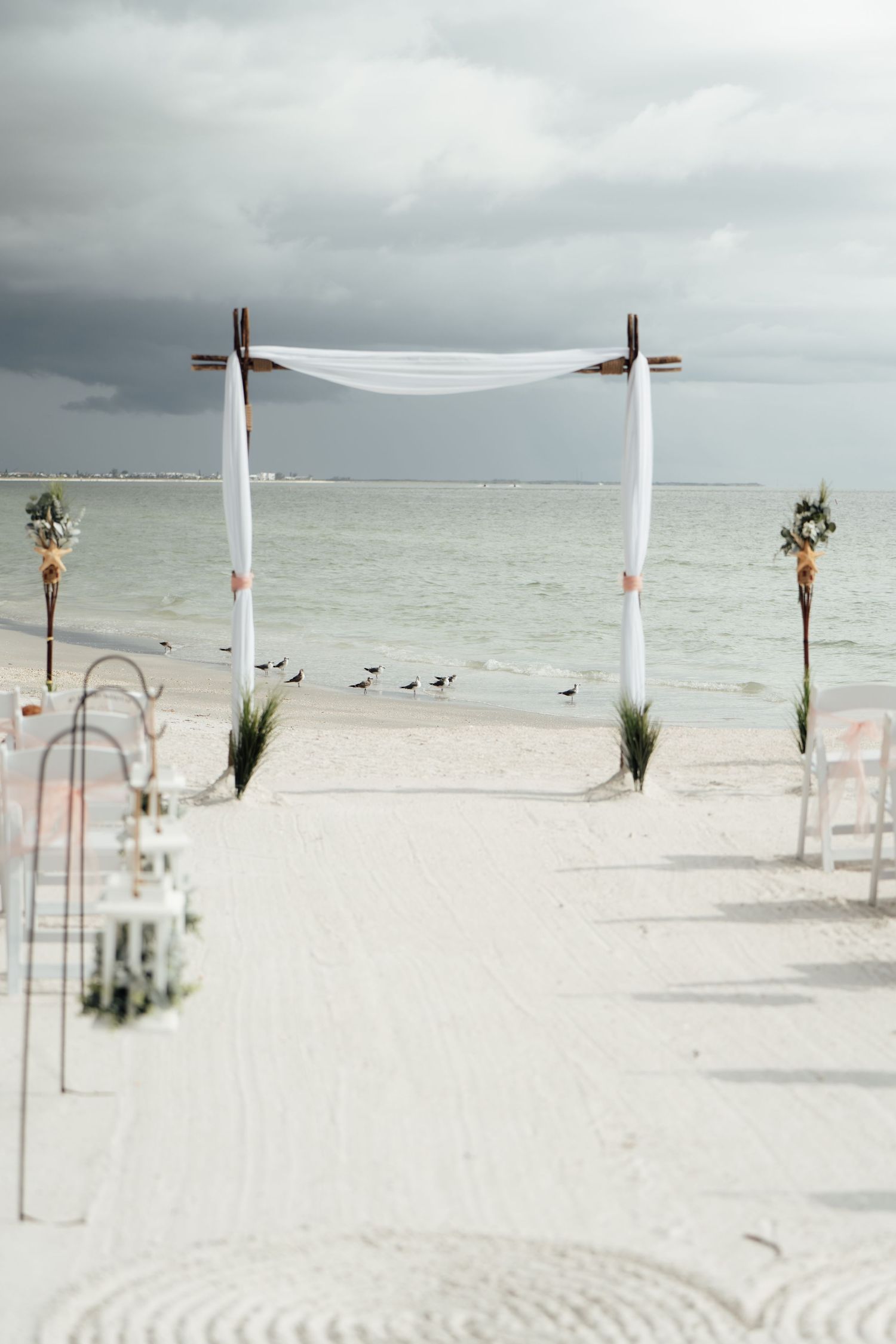 Beach wedding arch draped in white fabric on white sand, with ocean backdrop and seabirds on a cloudy day.