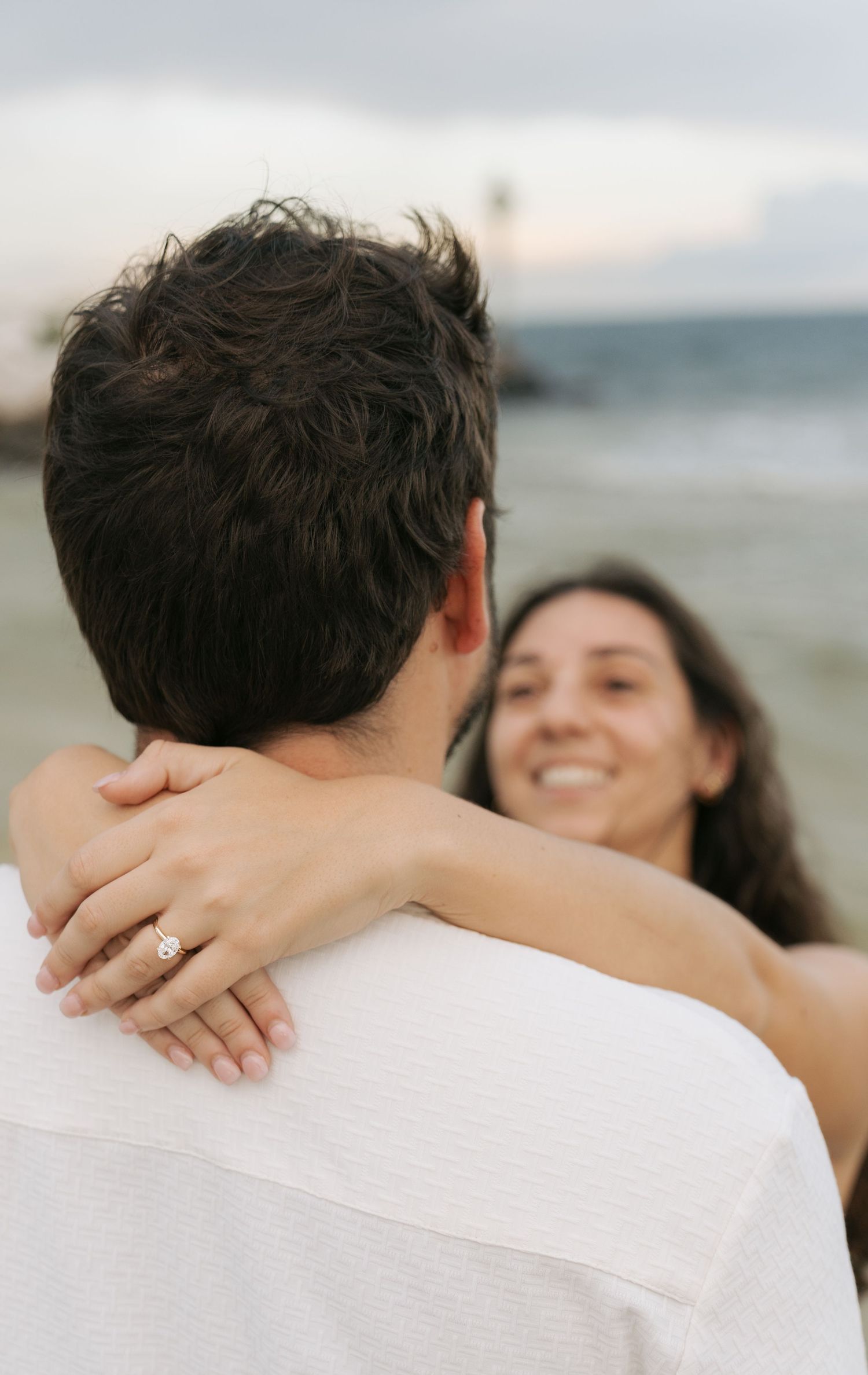A smiling woman with an engagement ring embraces a man on a beach at sunset.