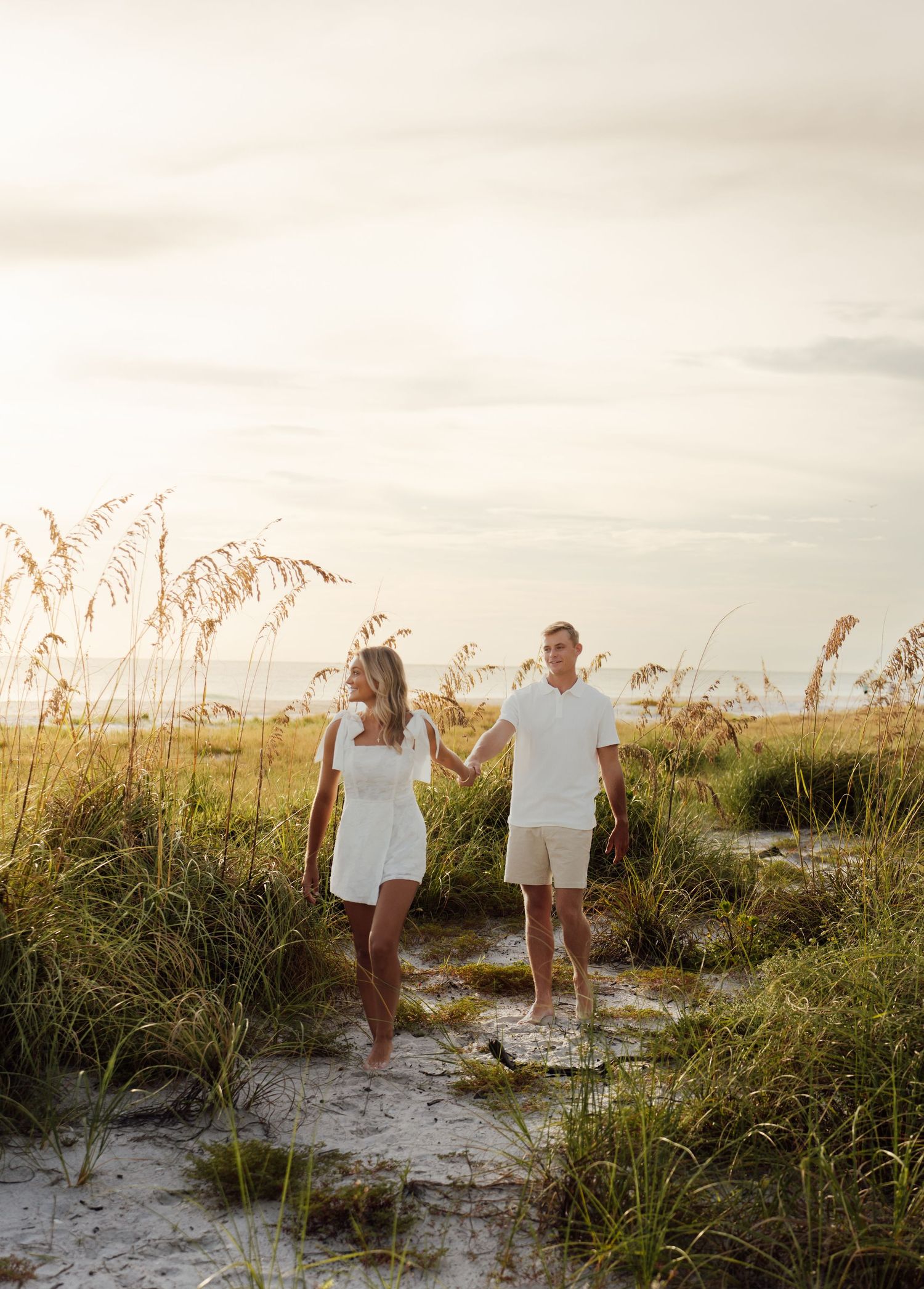 Couple in white outfits holding hands on a sandy beach path surrounded by sea oats at golden hour.