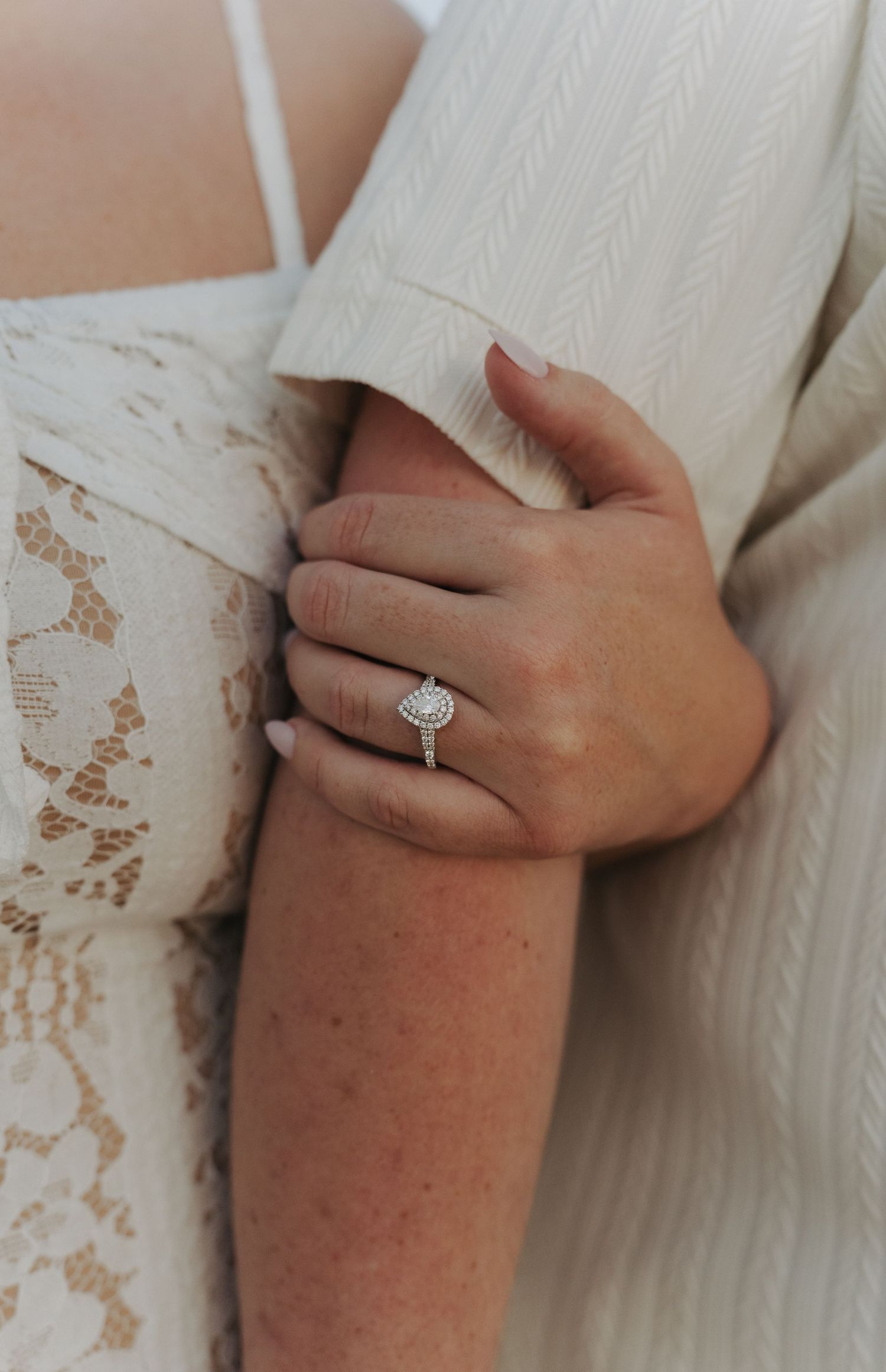 Close-up of a diamond halo engagement ring on a woman's finger, wearing a white lace dress.