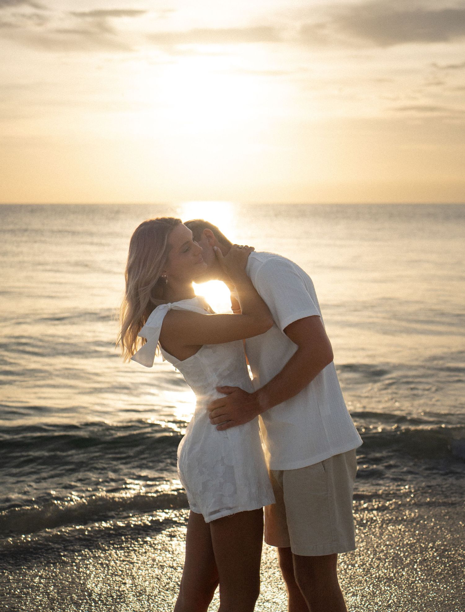Couple sharing a romantic kiss on the beach at golden sunset, wearing white outfits with ocean waves in the background.