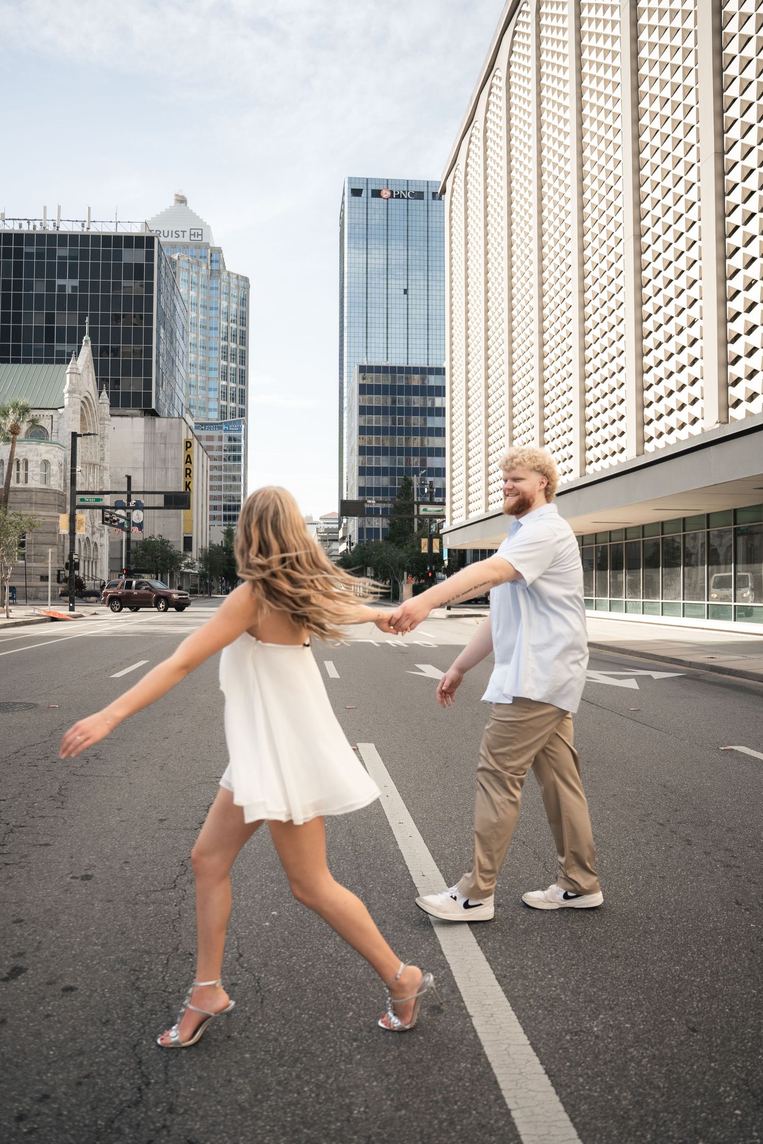 Couple holding hands dancing in urban street, woman in white dress, man in casual attire, city buildings backdrop.