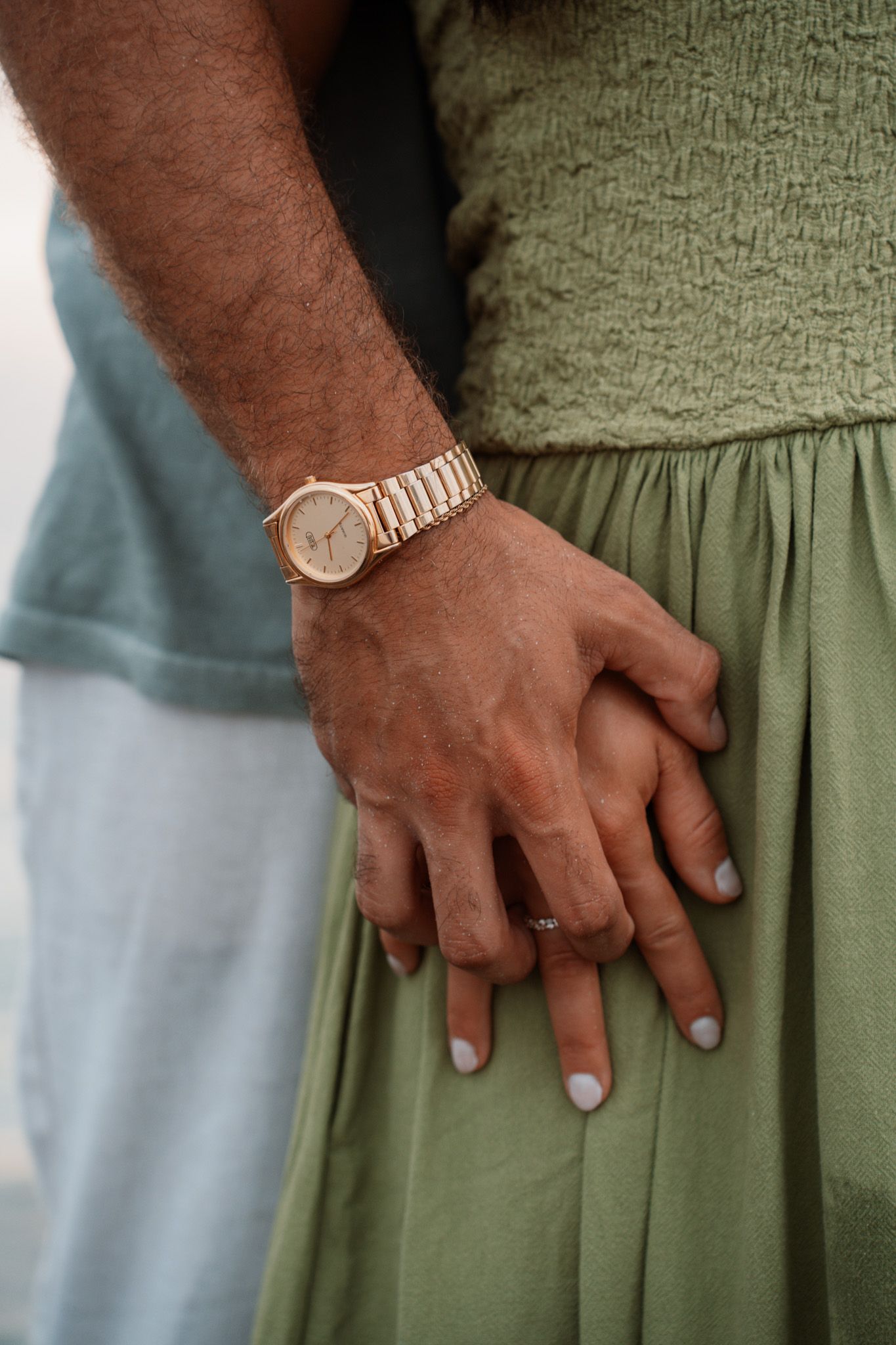 Close-up of couple holding hands, man wearing gold watch, woman in green dress with white manicure.