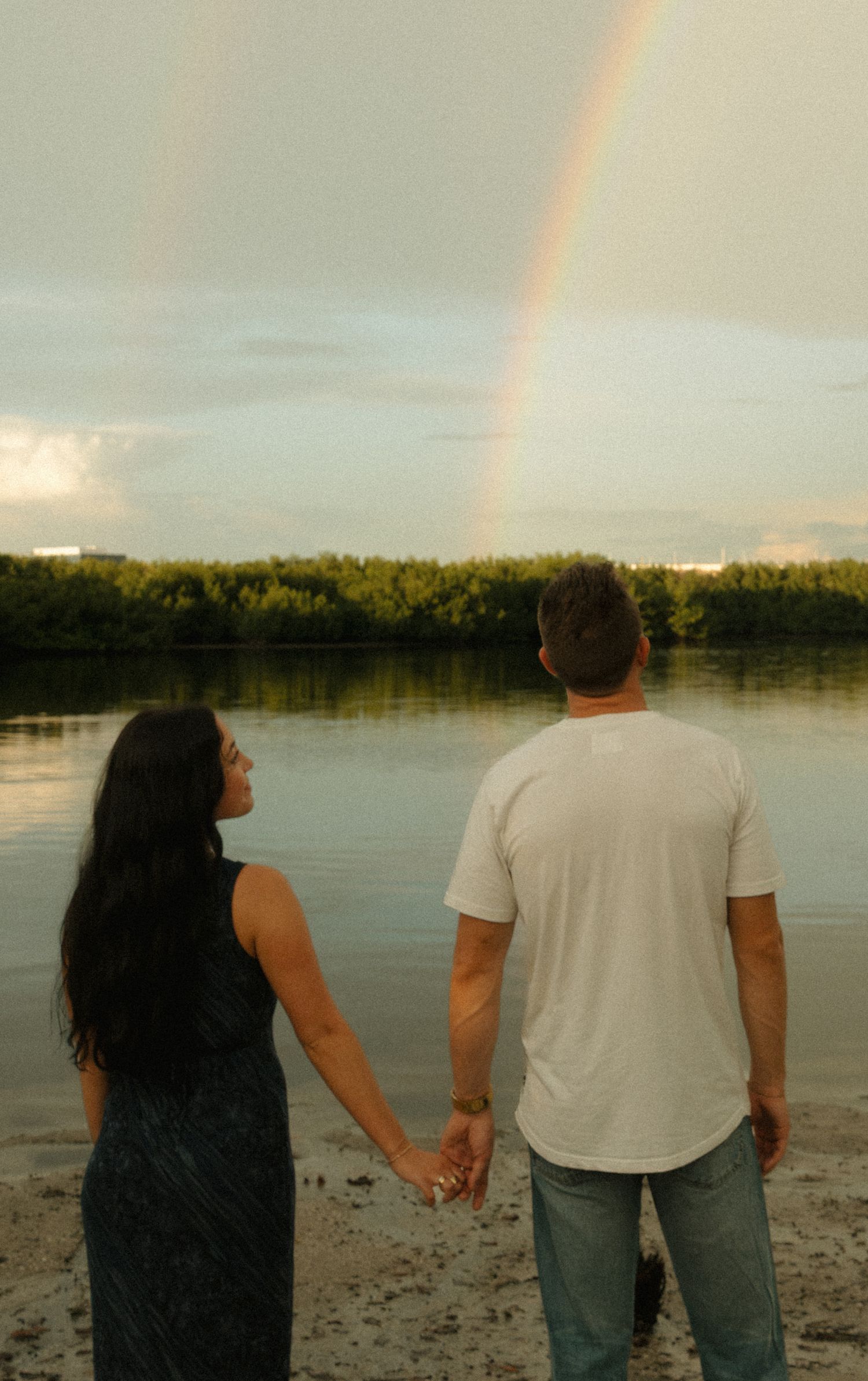 Couple holding hands on beach shore watching a rainbow over a tranquil river surrounded by lush green trees.
