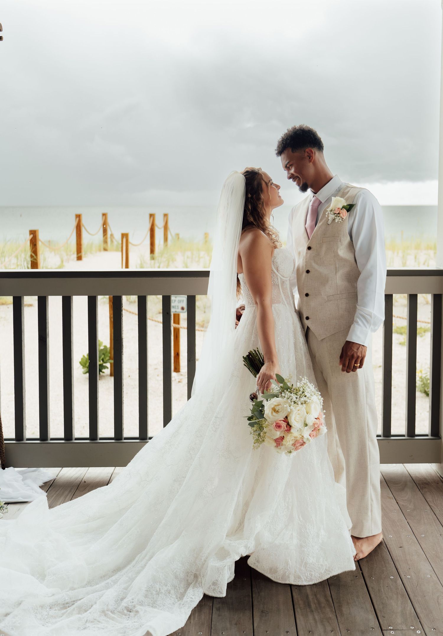 A bride and groom share an intimate moment on a waterfront deck, she in a flowing white gown holding a bouquet.