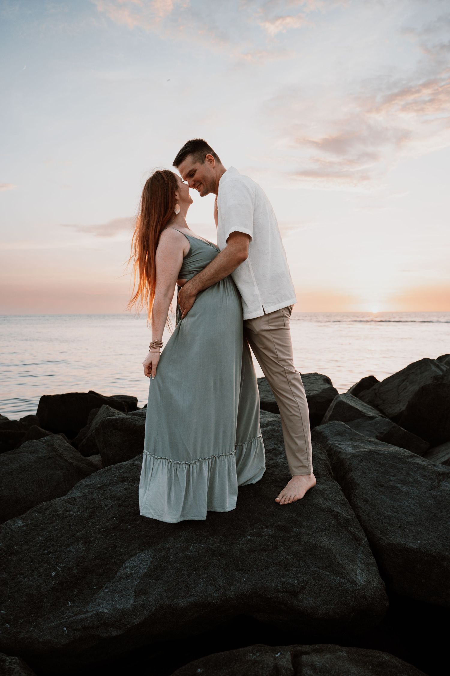 Couple sharing a romantic moment on rocky shoreline at sunset, woman in sage green maxi dress.