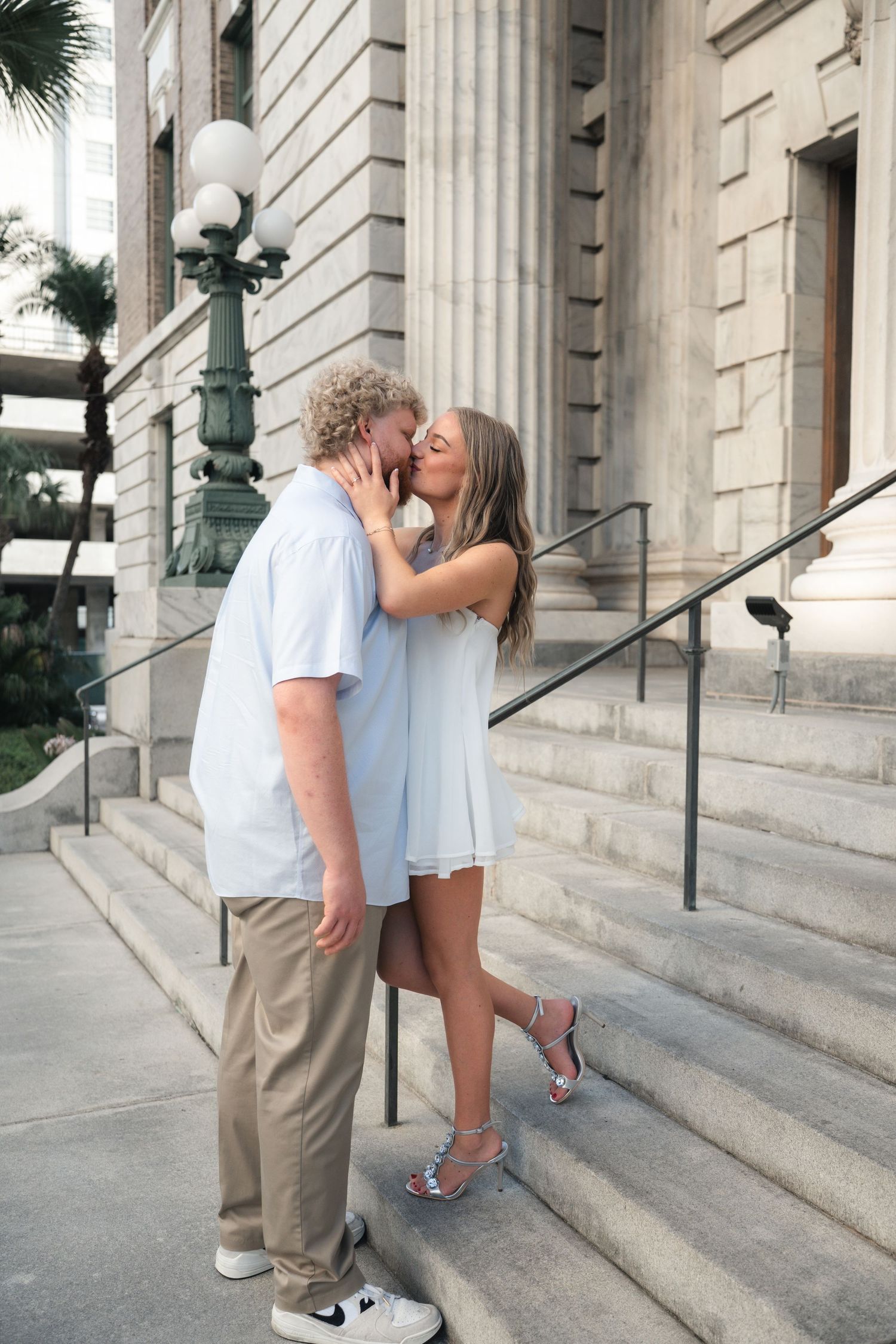 Couple sharing a romantic kiss on stone steps outside a classic building, woman in blue dress and heels.