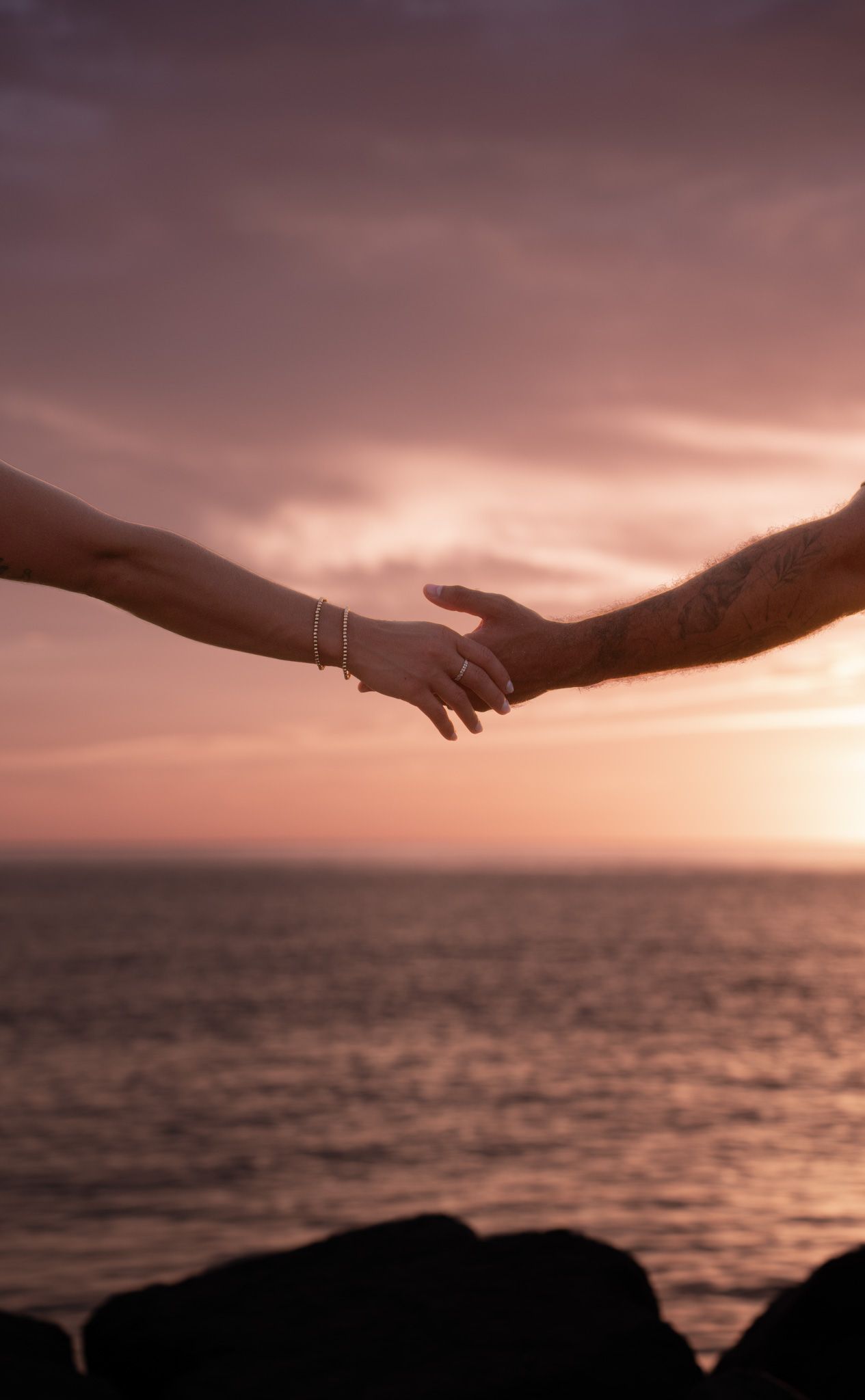 Two hands reaching toward each other at sunset over the ocean, one with a bracelet and ring, the other with a tattoo.