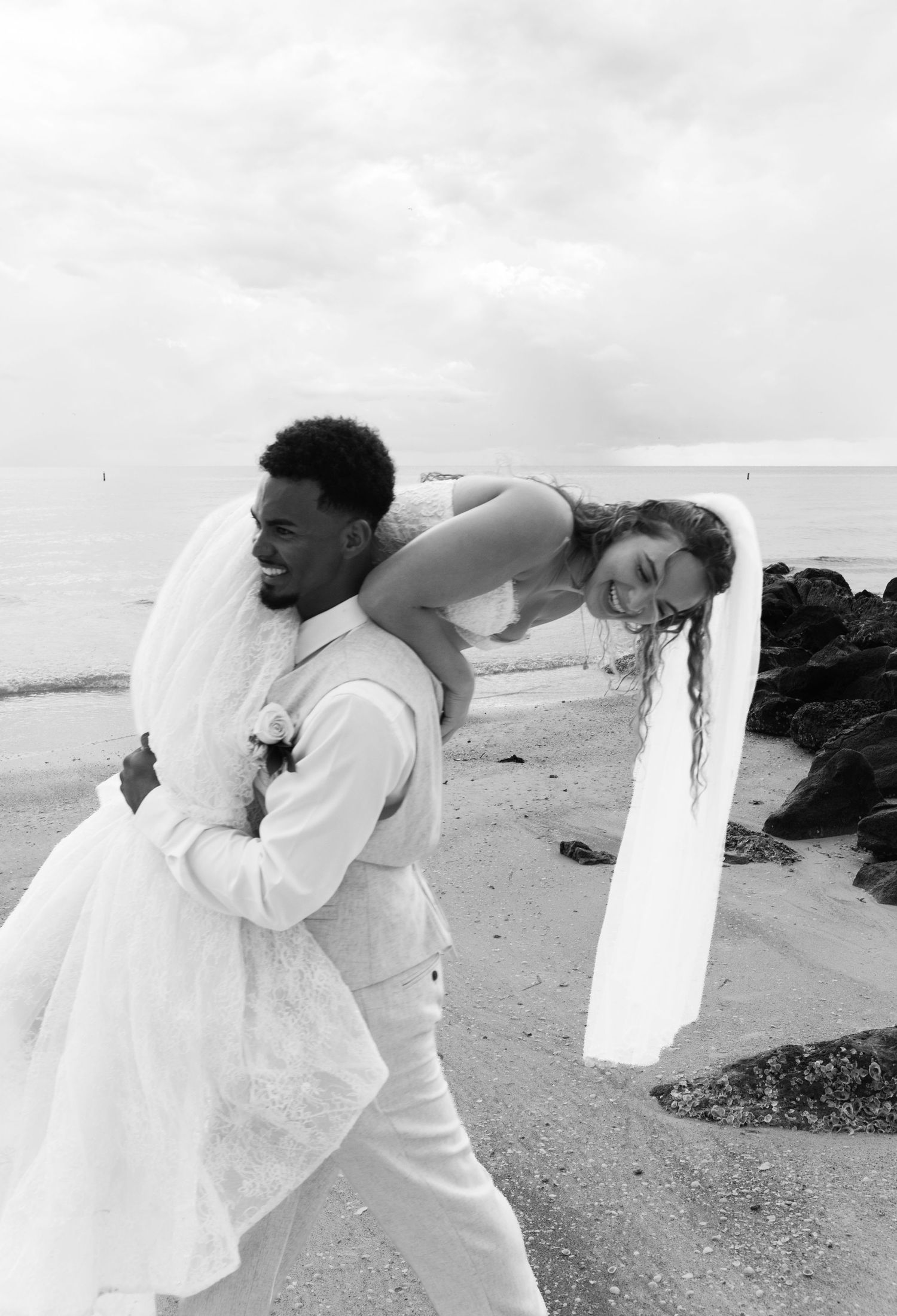 Black and white beach wedding photo of groom carrying laughing bride in veil and gown by rocky shoreline.