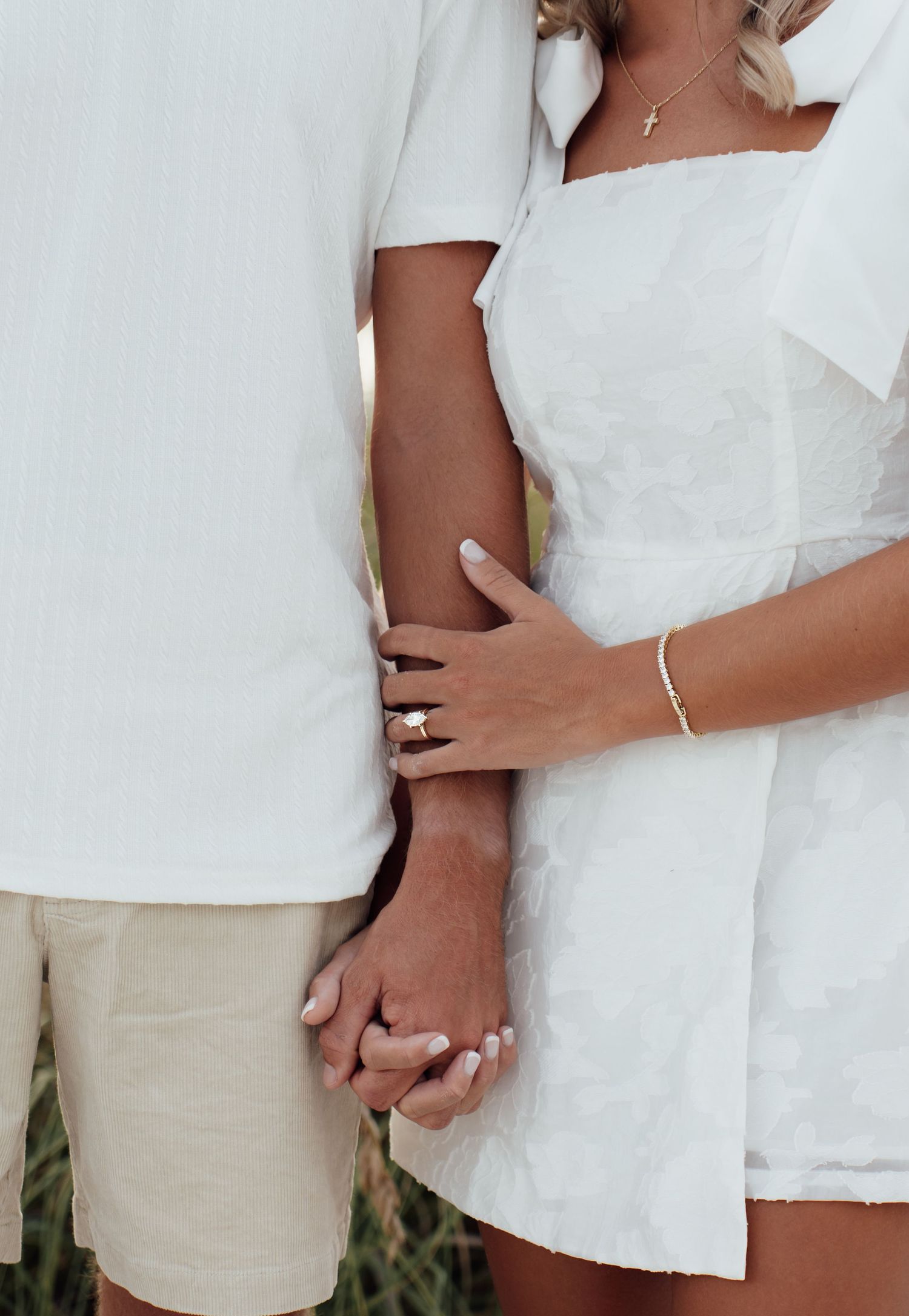 Couple in white outfits holding hands, woman wearing gold ring and bracelet with cross necklace.