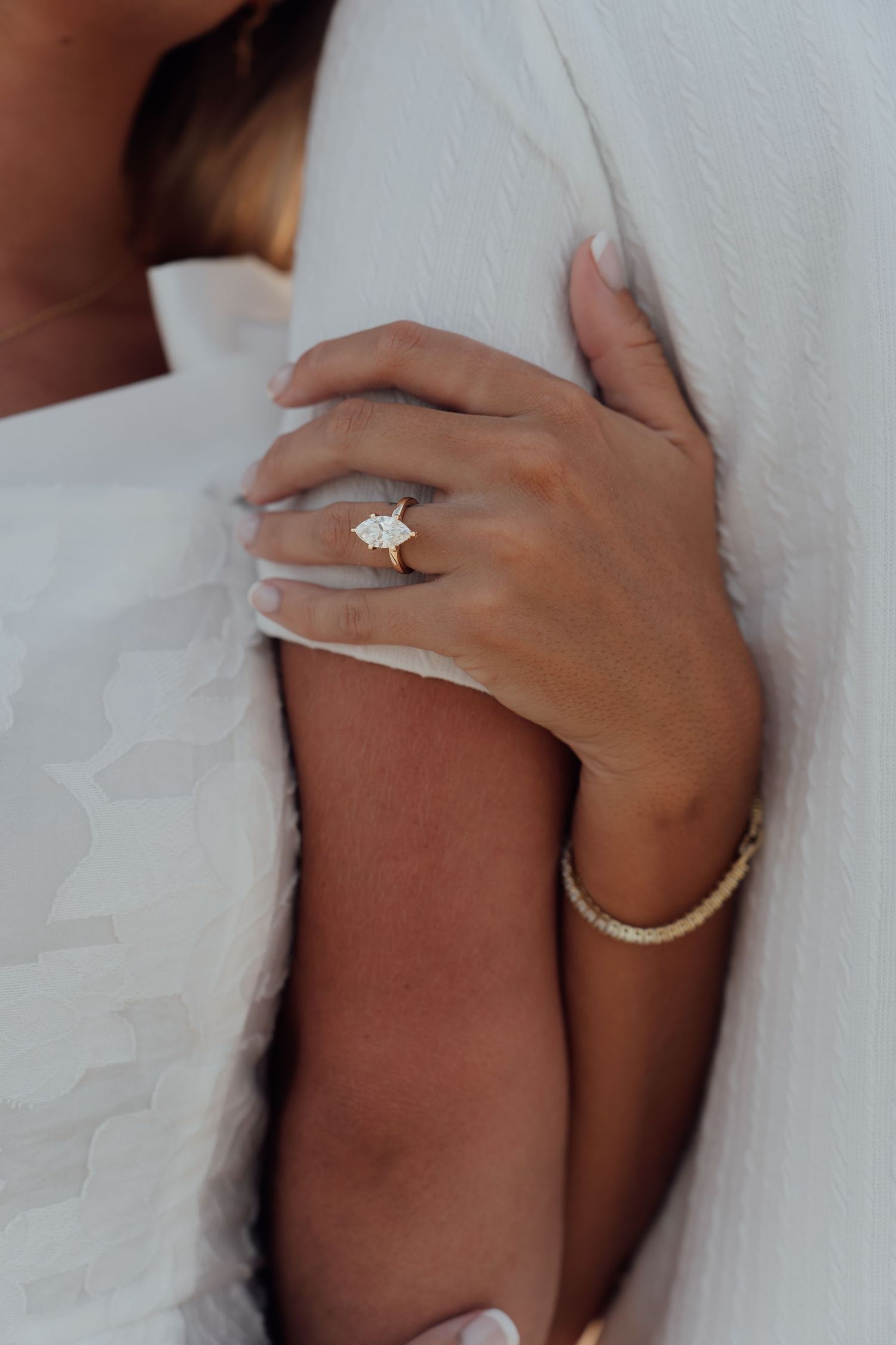 Close-up of a woman's hand wearing a gold diamond engagement ring and tennis bracelet on a white dress.