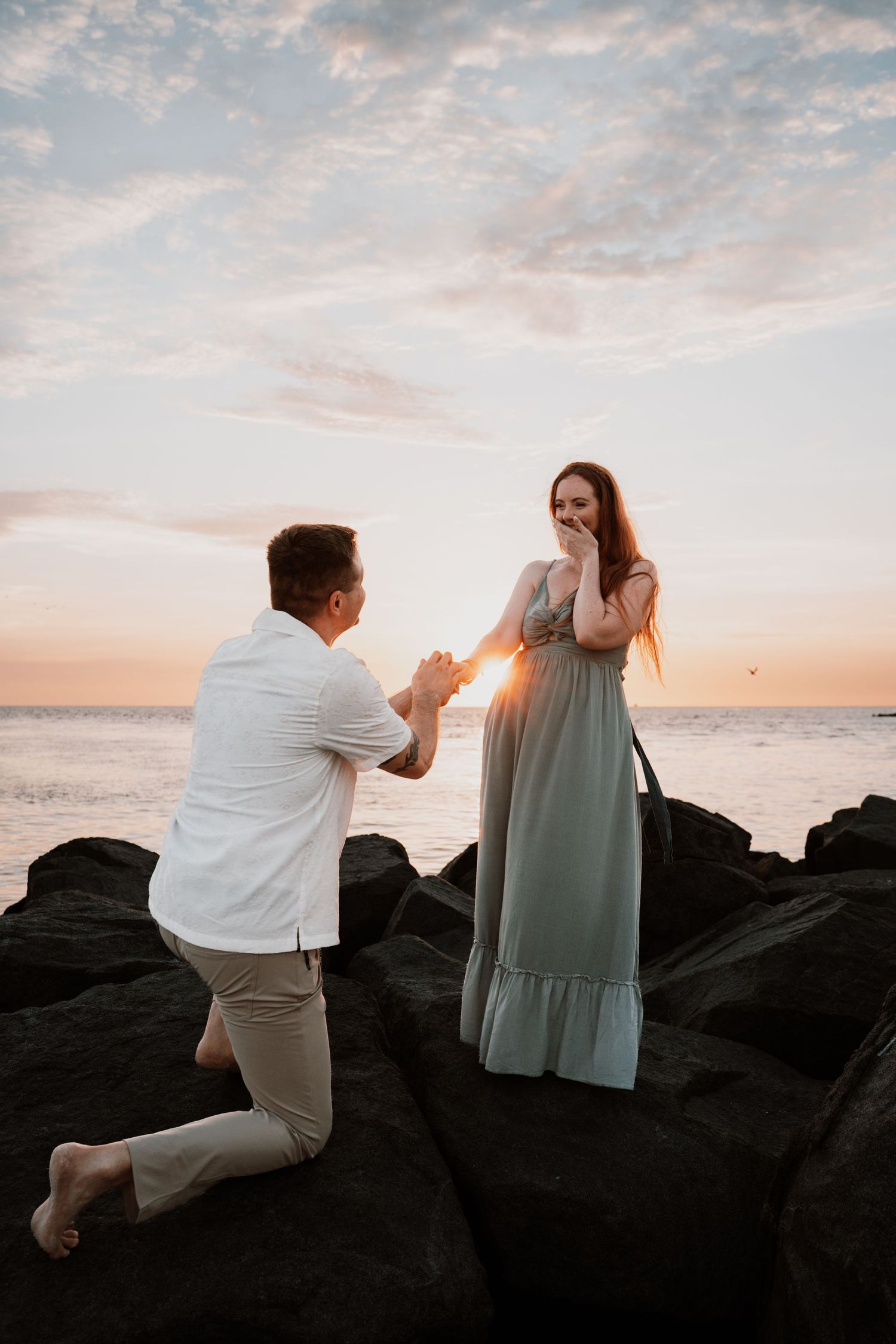 Man proposes to woman on rocky beach at sunset, she covers her mouth in surprise, wearing a sage green dress.