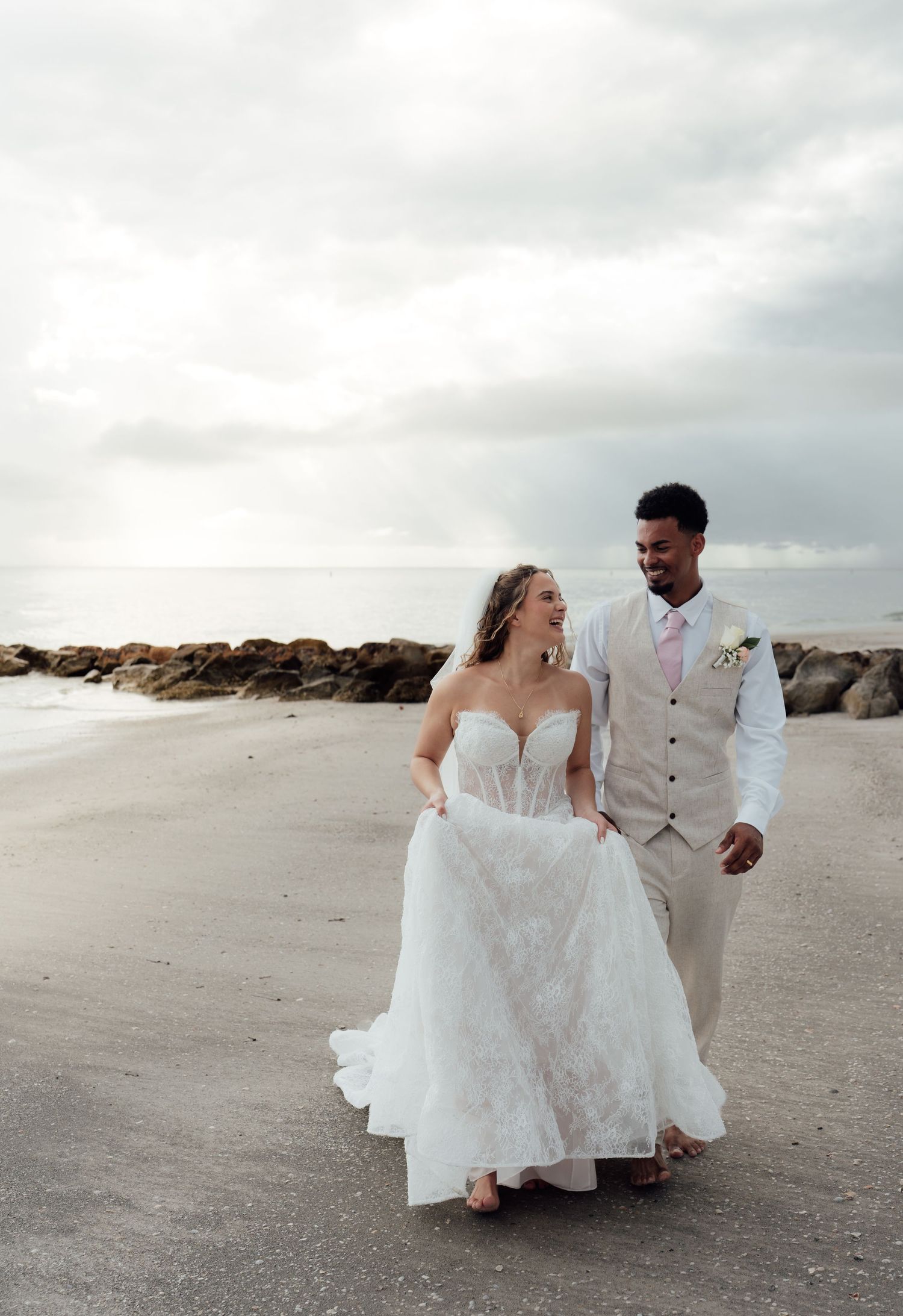 Bride and groom walk hand in hand on a sandy beach in wedding attire, with rocky shoreline and cloudy sky behind them.