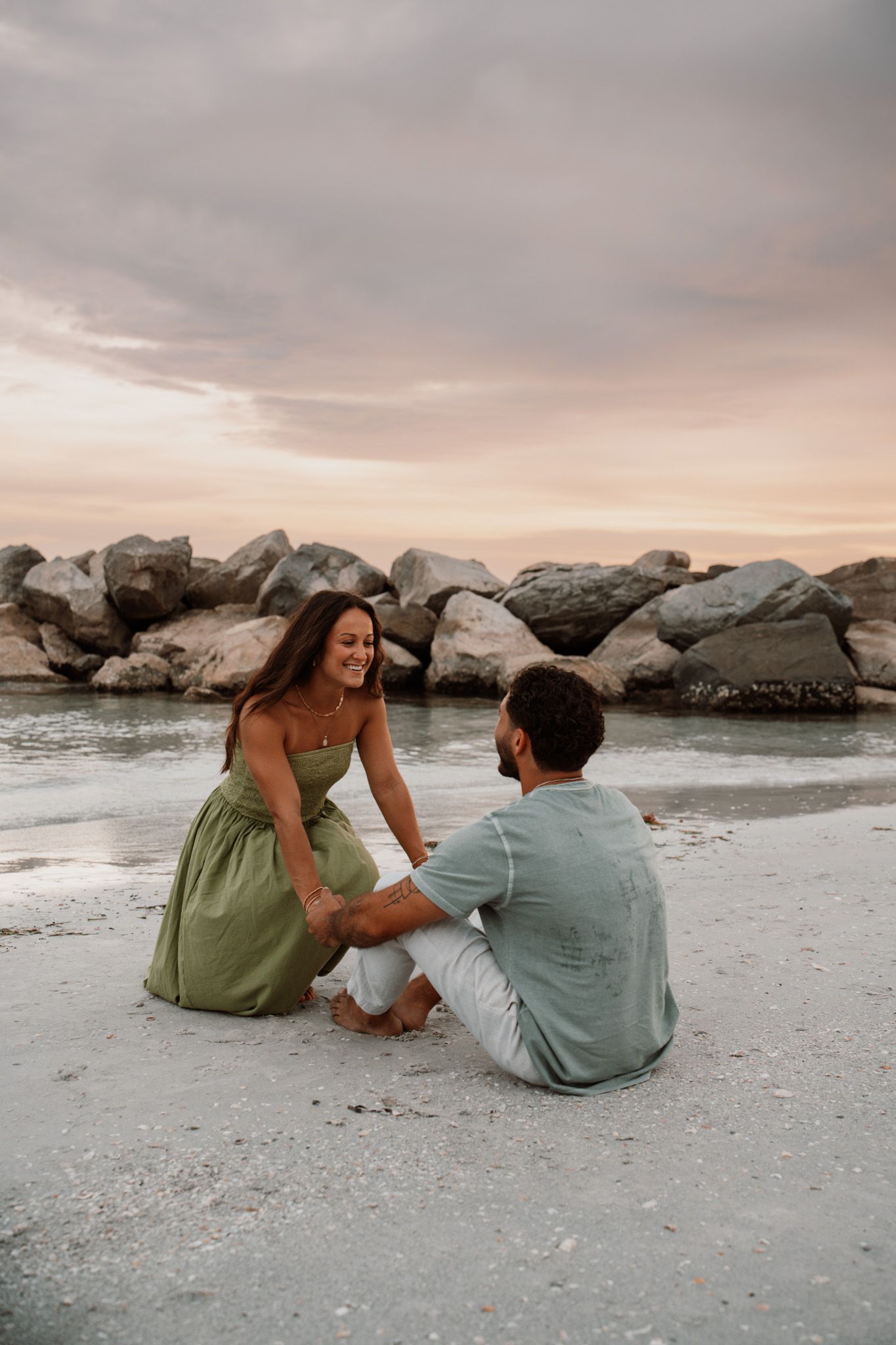 Couple sitting on sandy beach at sunset holding hands, woman in green dress smiling, rocks and colorful sky behind them.