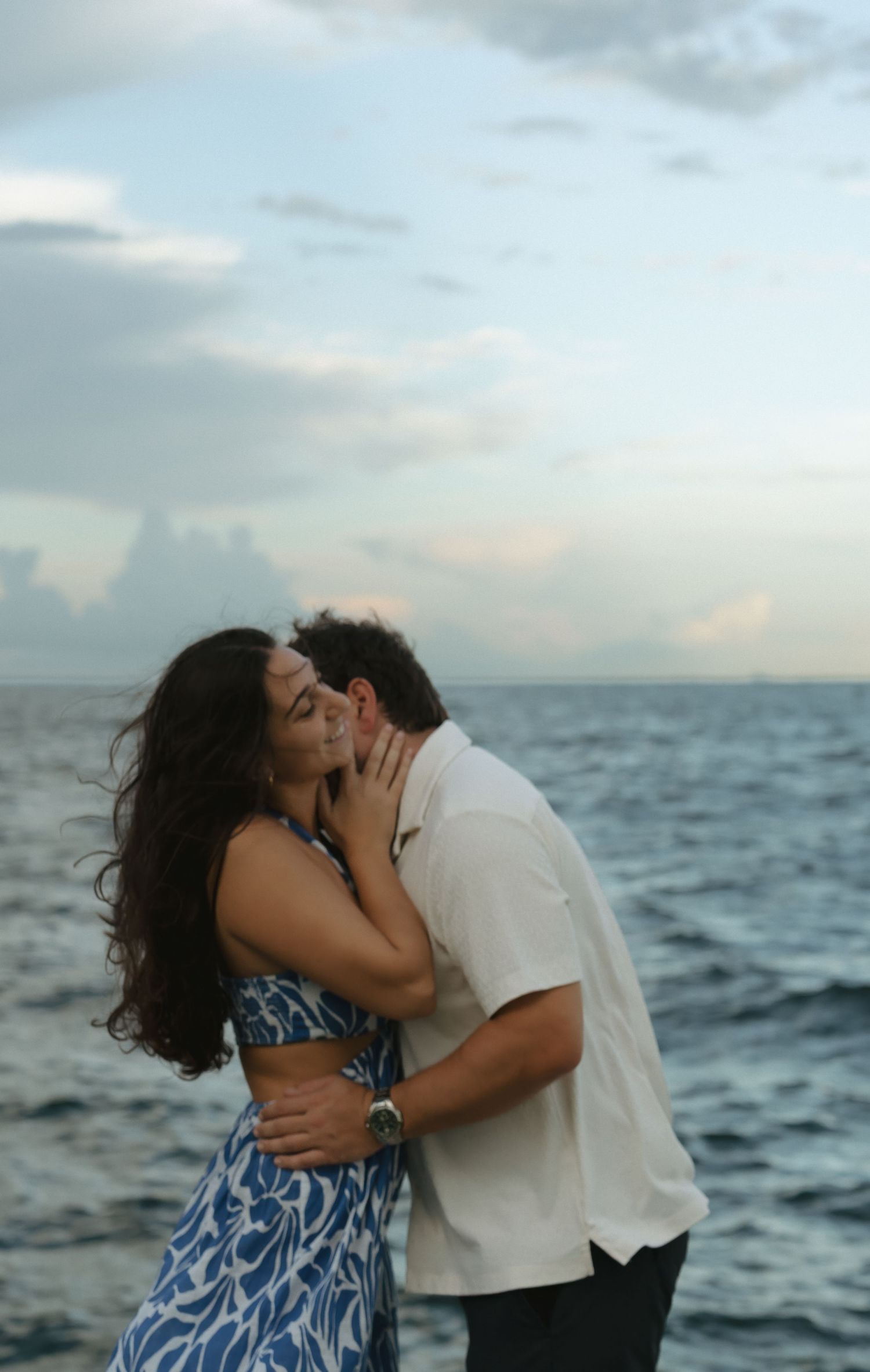 Couple sharing a romantic kiss on a seaside pier with ocean waves and cloudy sky in the background.
