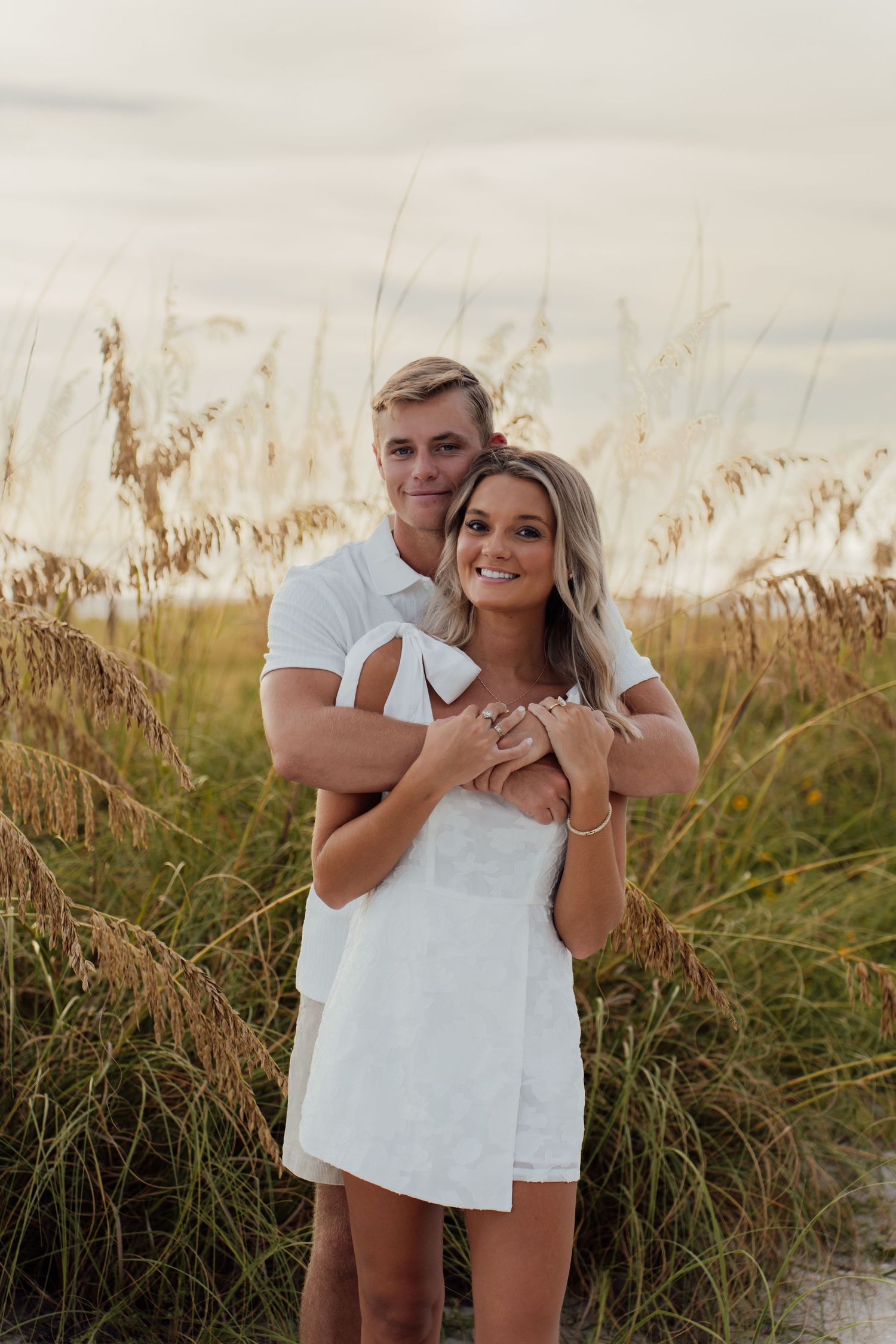 A smiling couple in white outfits embraces among tall sea oats grass during a golden hour beach photo session.