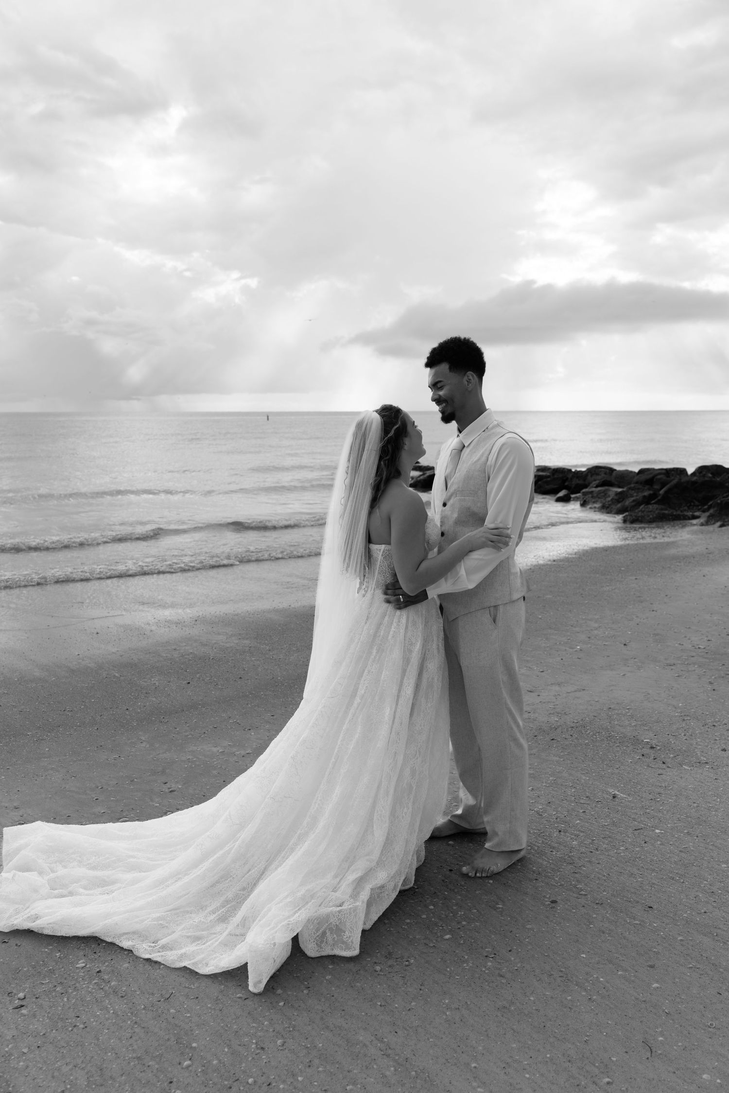 Black and white photo of a bride and groom embracing on a beach, with ocean waves and dramatic cloudy sky behind them.