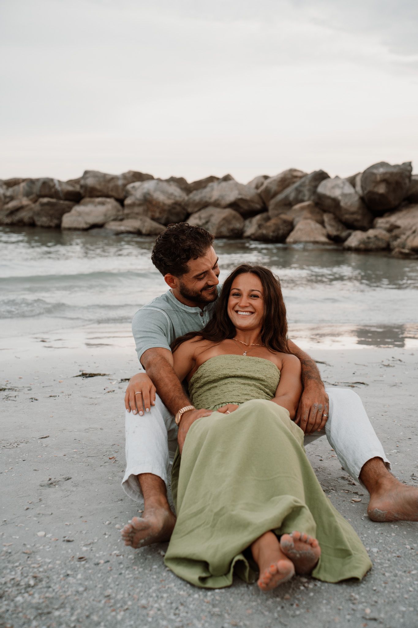 A couple sits together on a sandy beach, smiling, with rocky shoreline and calm ocean waves in the background.