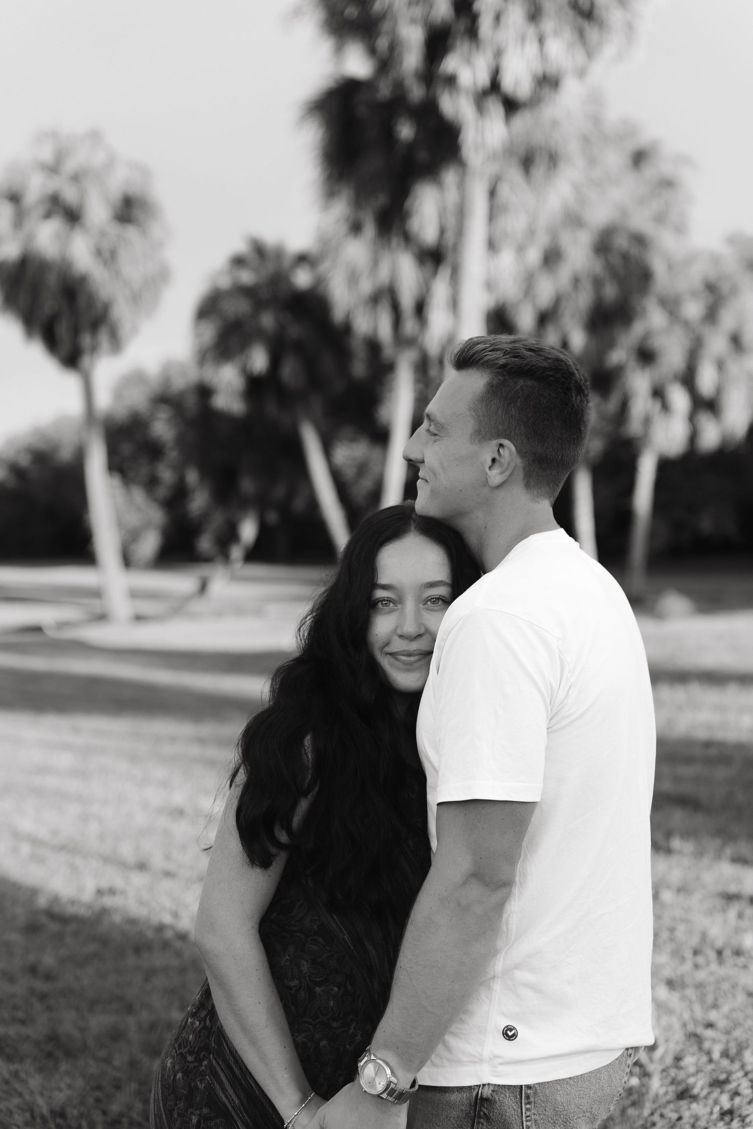 Black and white photo of a couple embracing outdoors with palm trees, woman smiling warmly at camera.