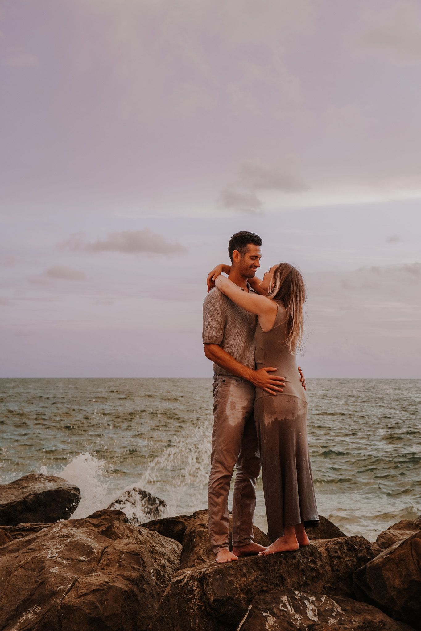 Couple embracing on rocky shoreline at sunset, waves crashing behind them near the ocean.