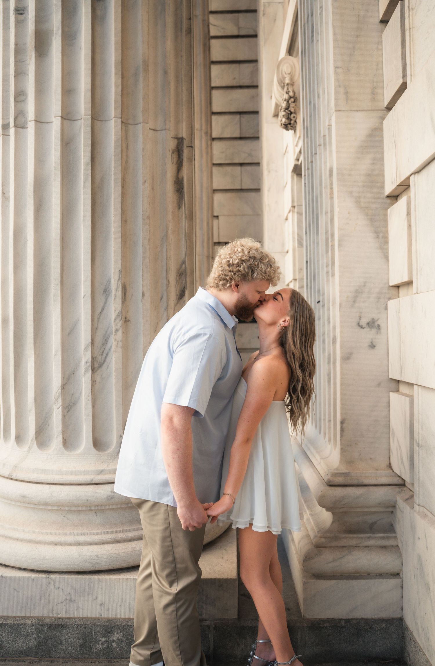 A couple shares a romantic kiss between marble columns of a neoclassical building, she in a white dress, he in a blue shirt.