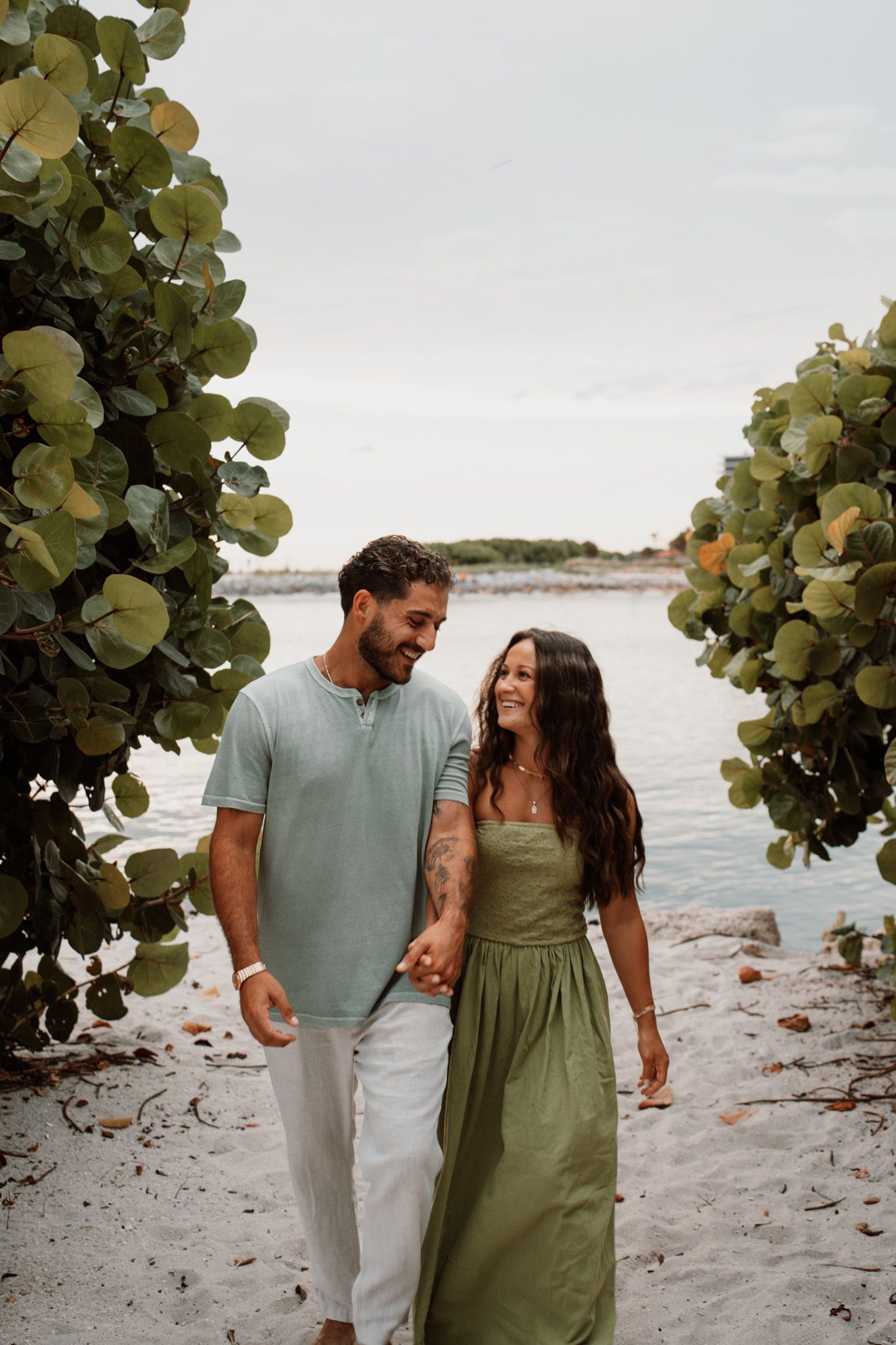 A smiling couple holds hands walking on a sandy beach path framed by tropical trees near calm water.